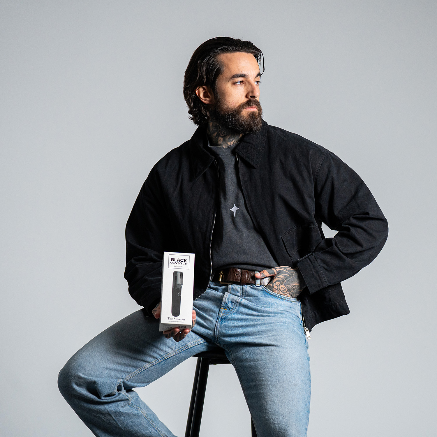 Uomo tatuato che tiene una scatola di prodotto. Indossa una giacca nera e jeans blu, seduto su uno sgabello. Lo sfondo è grigio.