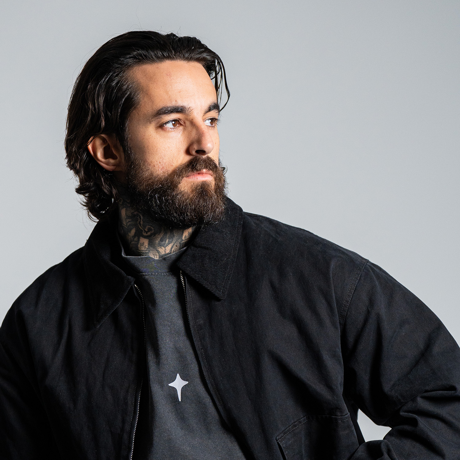 Un hombre con barba y tatuajes mira hacia un lado. Lleva una chaqueta negra sobre una camisa gris con un logotipo de estrella.