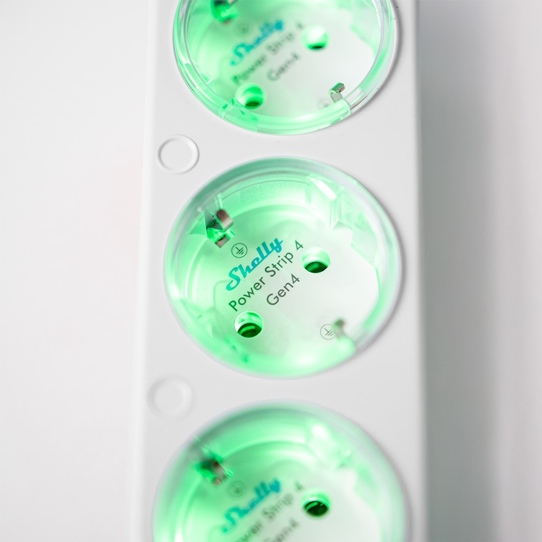 Zbliżenie białej listwy zasilającej z trzema podświetlanymi zielonymi gniazdami. Każde gniazdo ma markę 'Shelly'.