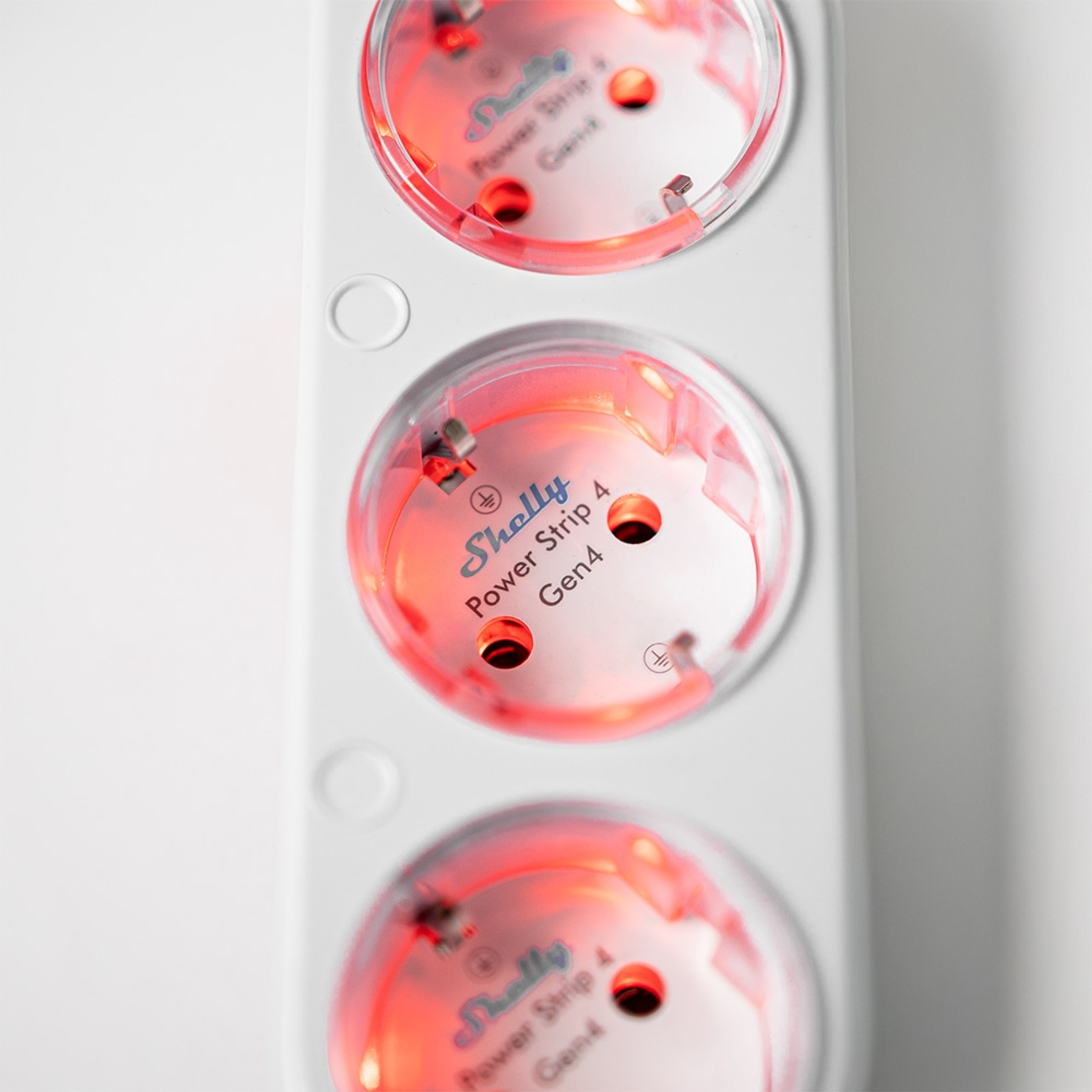 Zbliżenie białej listwy zasilającej z trzema gniazdami, każde podświetlone czerwonym światłem i logo Shelly.