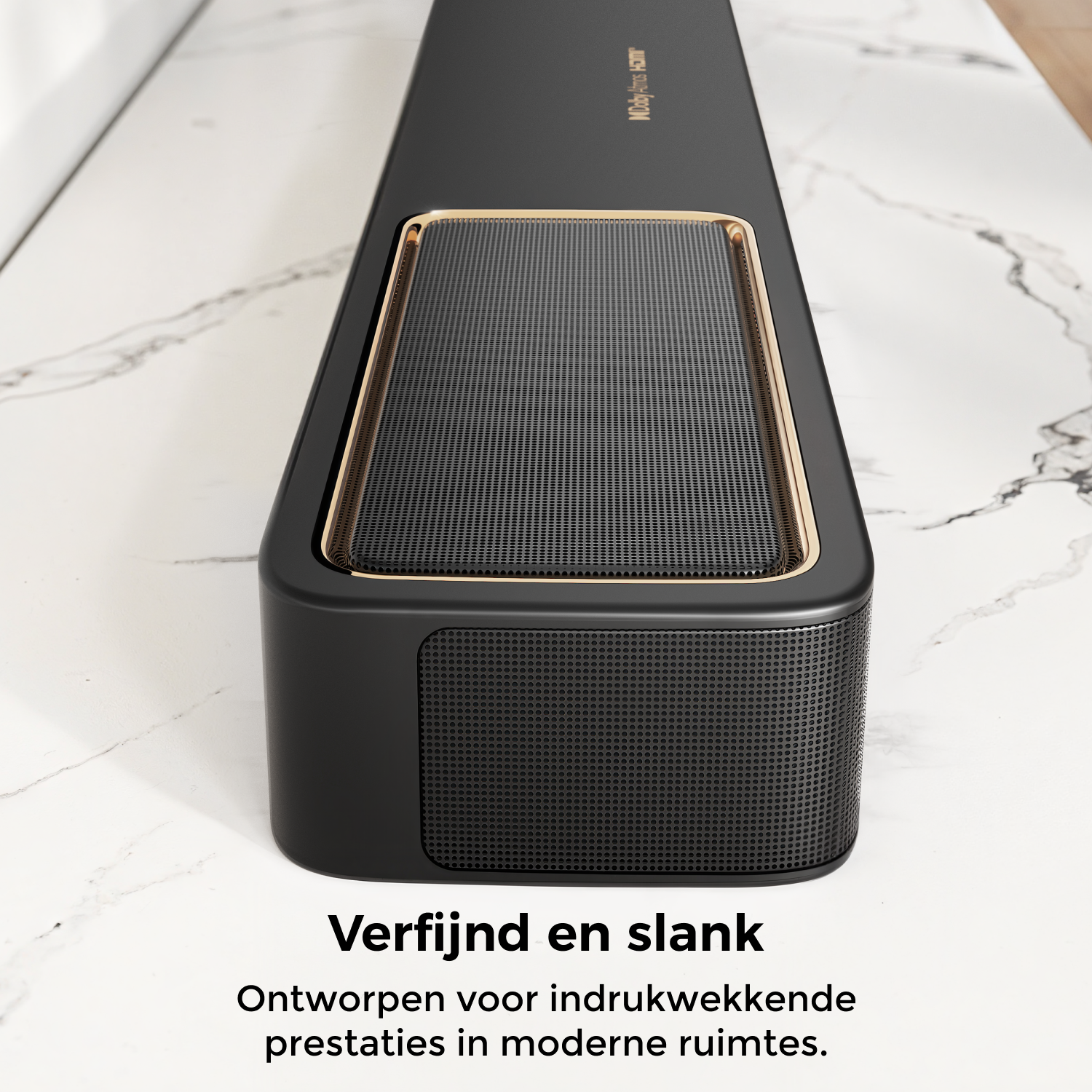 A sleek black soundbar with a gold trim sits on a marble surface. The soundbar has a mesh front and top, with text below.