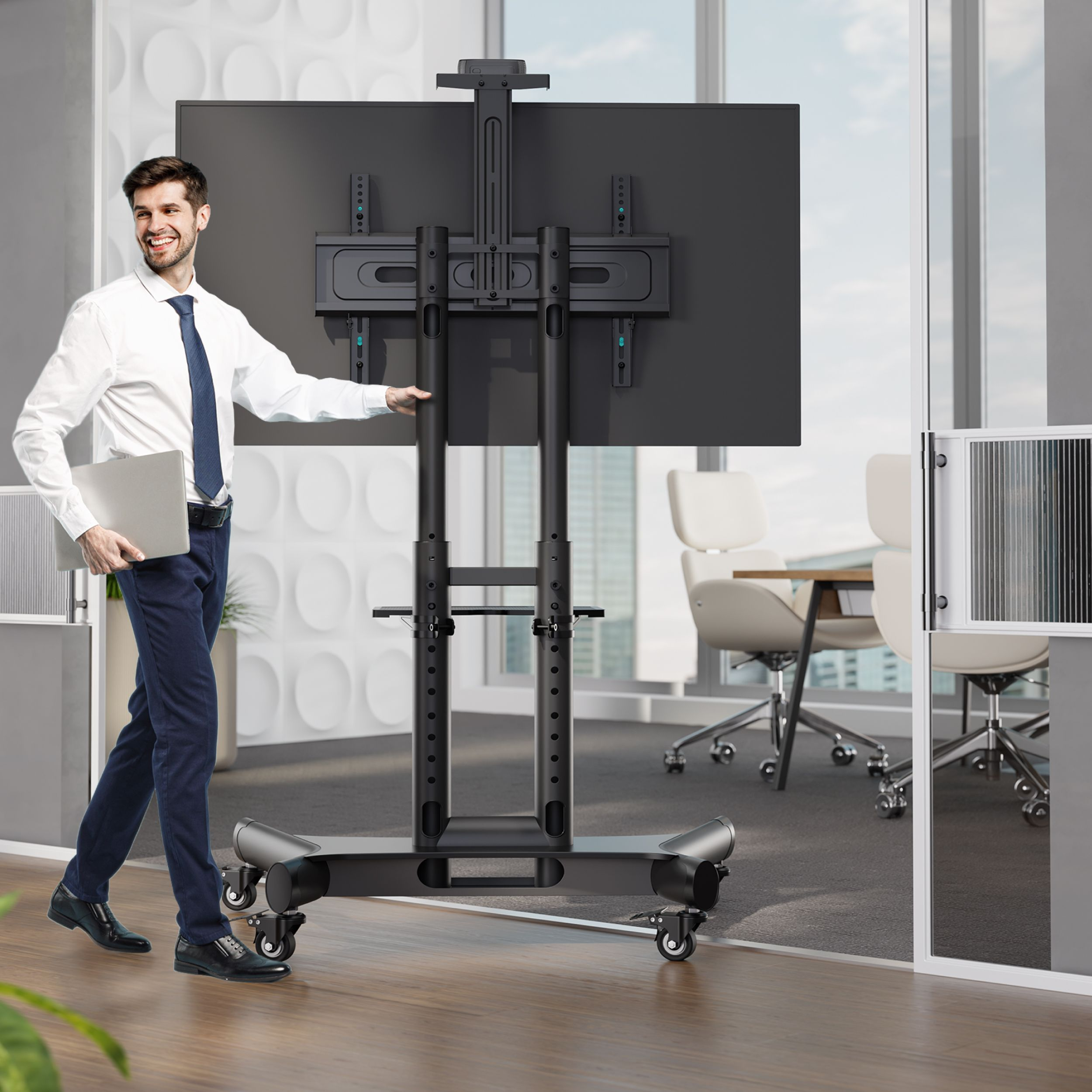 Mężczyzna idzie w biurze trzymając laptopa. Za nim duży telewizor na stojaku na kółkach i sala konferencyjna.