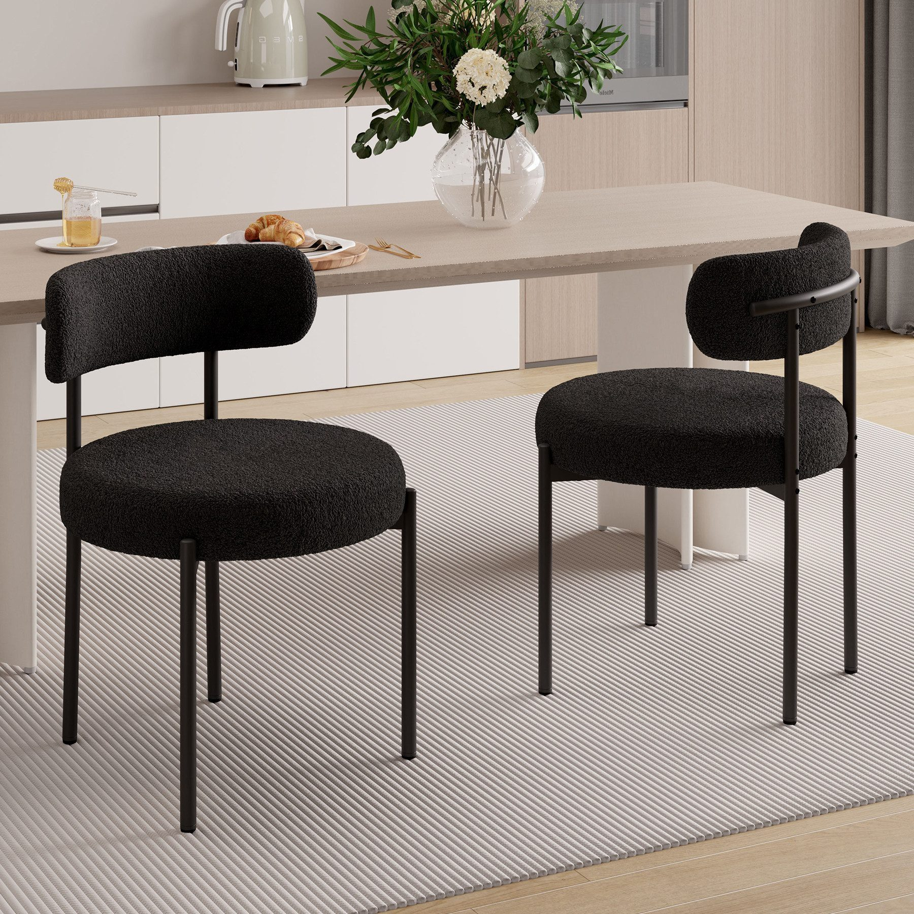 Zwei schwarze Stühle mit strukturierten Sitzen stehen auf einem gestreiften Teppich in der Nähe eines Tisches mit Blumen. Der Hintergrund beinhaltet eine Küche.