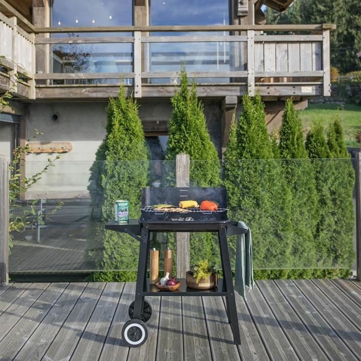 Een zwarte BBQ met eten op een houten terras voor een huis. Groene bomen en een glazen hek op de achtergrond.