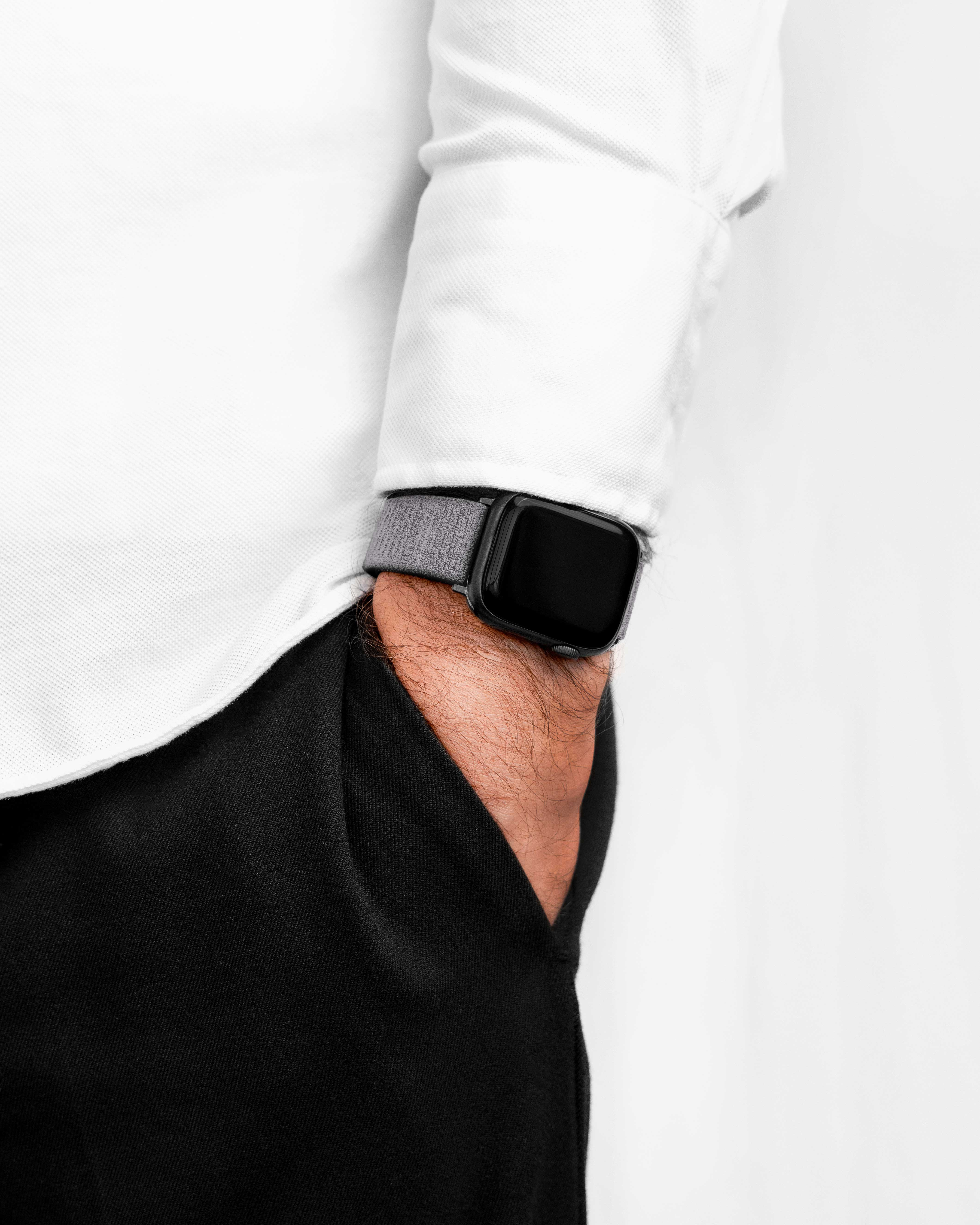 Der Arm einer Person mit einer grauen Uhr am Handgelenk, die Hand in der Tasche. Sie trägt ein weißes Hemd und eine schwarze Hose.