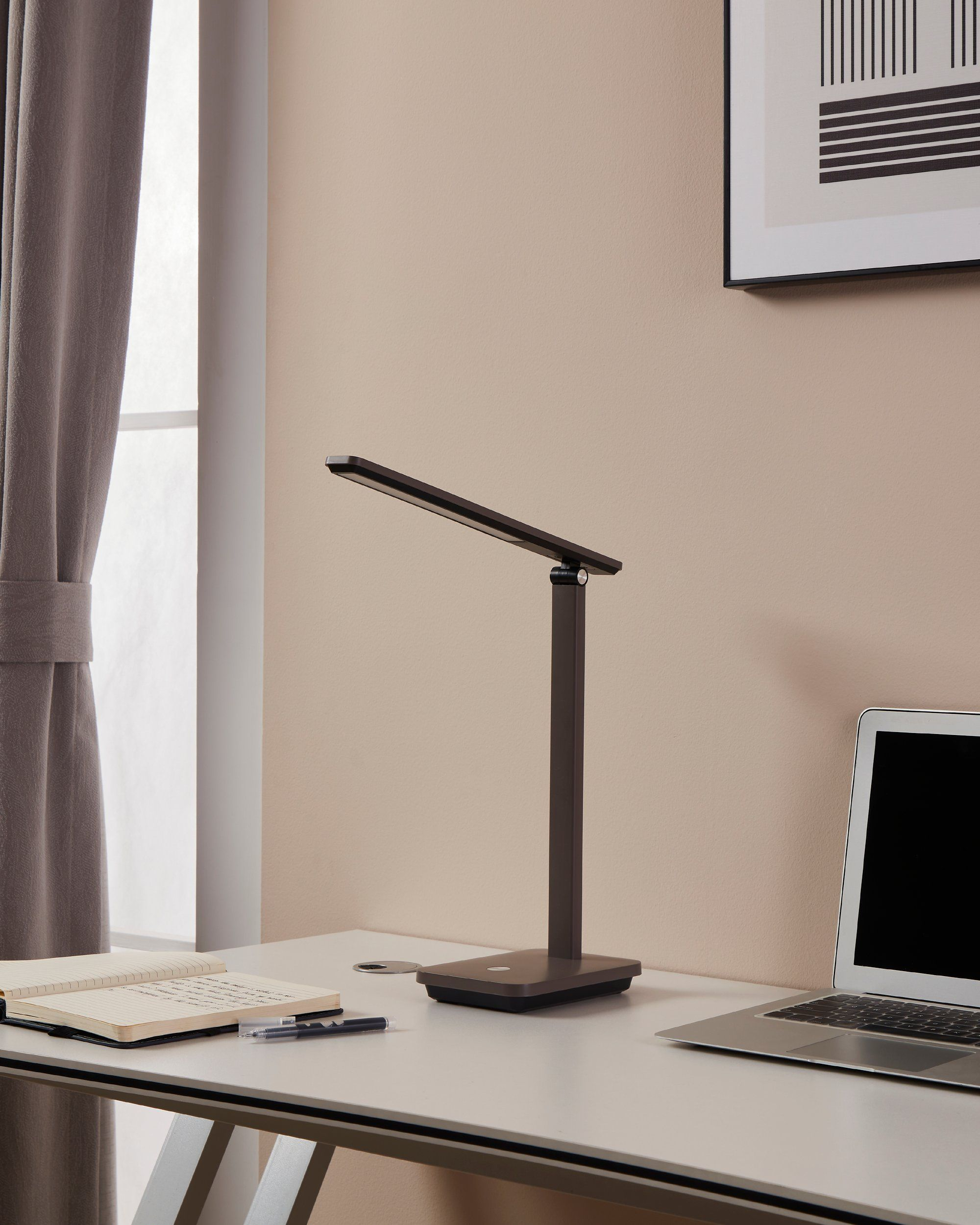 Ein Schreibtisch mit Lampe, Notizbuch, Stift und Laptop. Ein Fenster und Kunstwerk im Hintergrund.