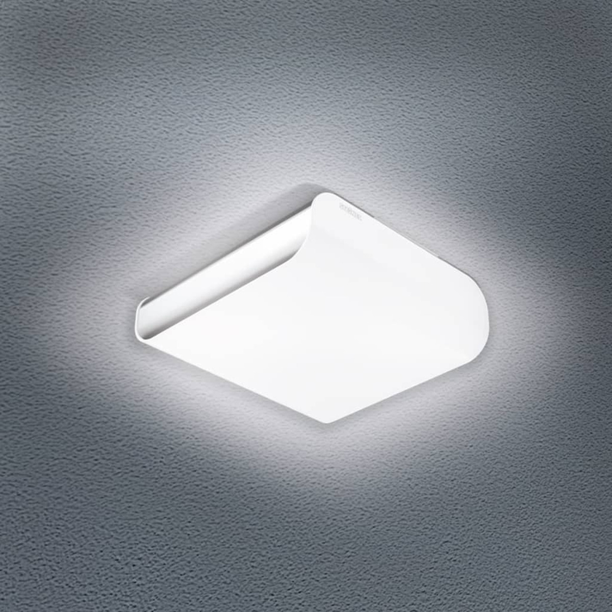 Un plafonnier blanc et carré est centré dans le cadre. La lumière est allumée, illuminant le plafond gris texturé en dessous.