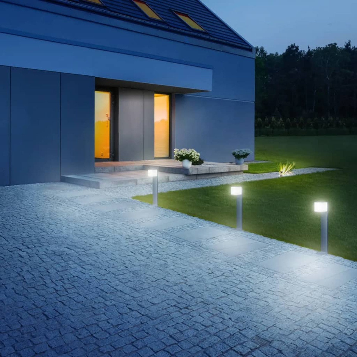 Außenansicht eines modernen Hauses in der Dämmerung. Beleuchteter Weg führt zur Tür. Rasen und Bäume im Hintergrund. Lichter sind an.