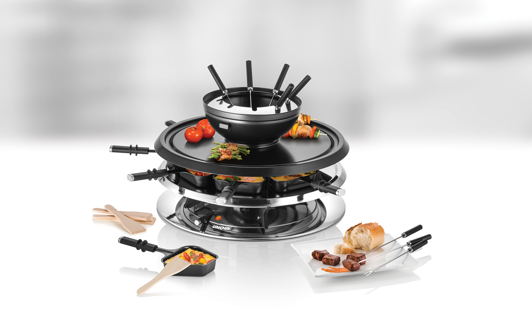 Ein Raclette-Grill mit Lebensmitteln, darunter Tomaten, Spieße und Brot. Schwarzer Grill, silberner Sockel und kleine Pfannen. Weißer Hintergrund.