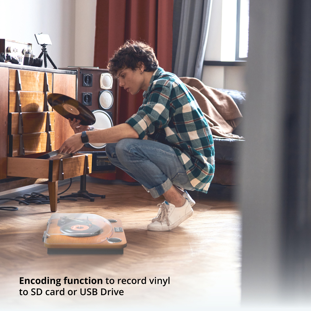 Mężczyzna w koszuli w kratę kuca obok gramofonu, trzymając płytę winylową. Drewniana szafka i wieża stereo są w tle.