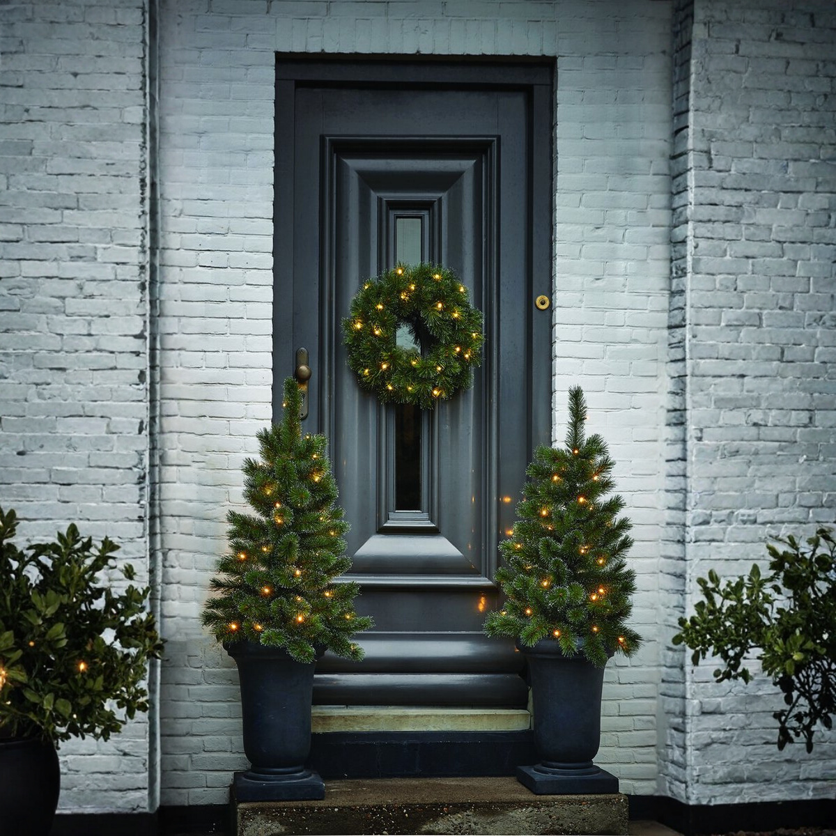 Une porte sombre avec une couronne et deux petits arbres de Noël en pots. Un mur de briques blanches des deux côtés de la porte.