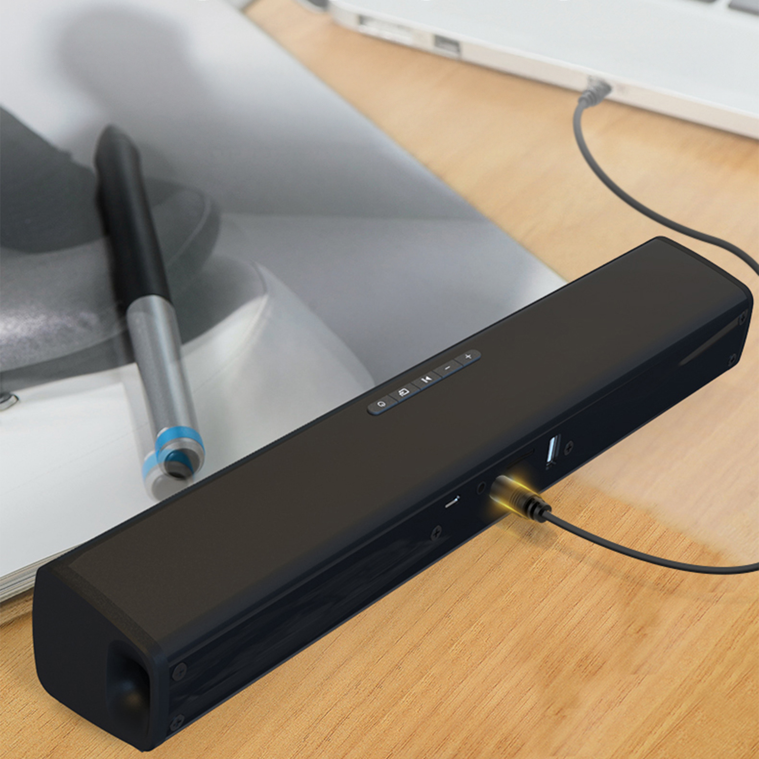 Soundbar nera su una scrivania di legno con una penna e un laptop sullo sfondo. Un cavo è collegato.