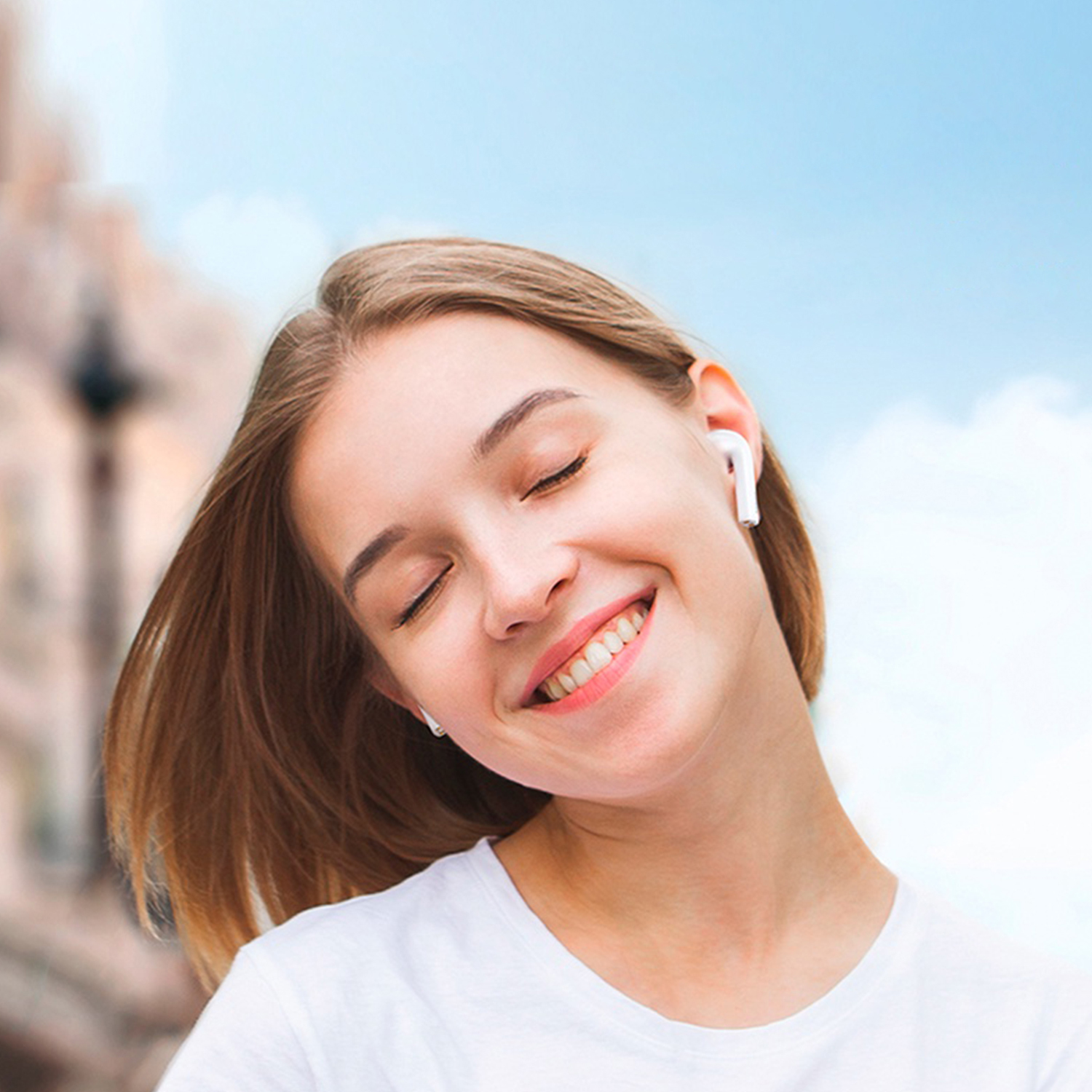 Una donna sorridente con gli occhi chiusi che indossa auricolari e una maglietta bianca. Lo sfondo mostra un edificio sfocato e cielo blu.