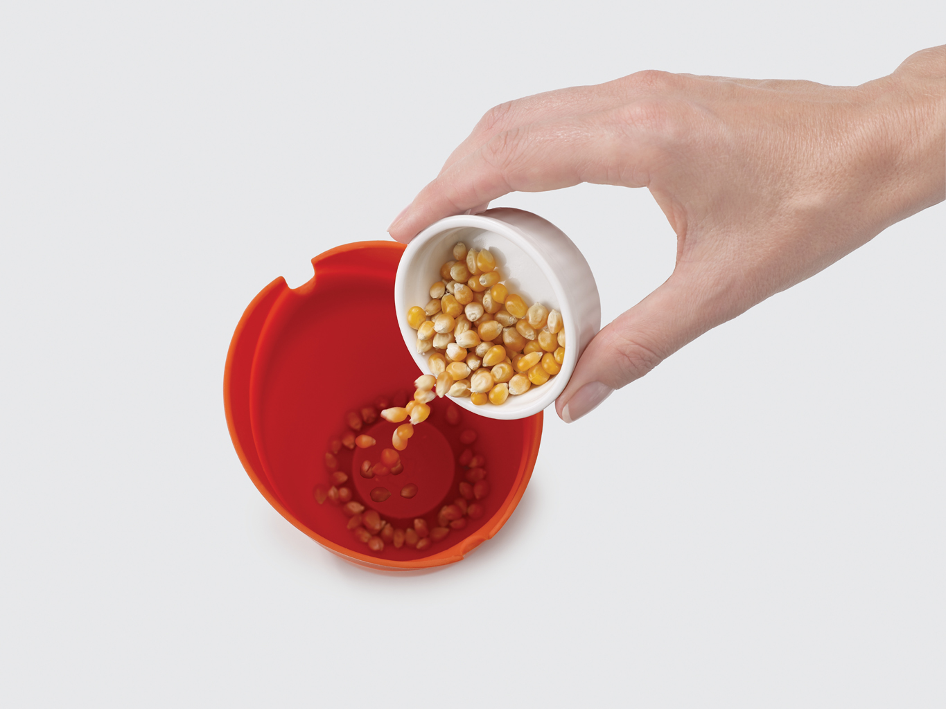 Una mano vierte granos de maíz de un cuenco blanco pequeño a una máquina roja para hacer palomitas. Los granos están dispersos dentro y fuera.