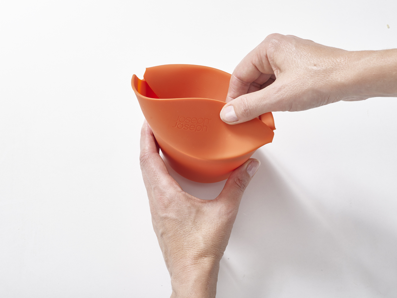 Manos sosteniendo un cuenco de silicona naranja. El cuenco tiene la marca 'Joseph Joseph'. El fondo es blanco.