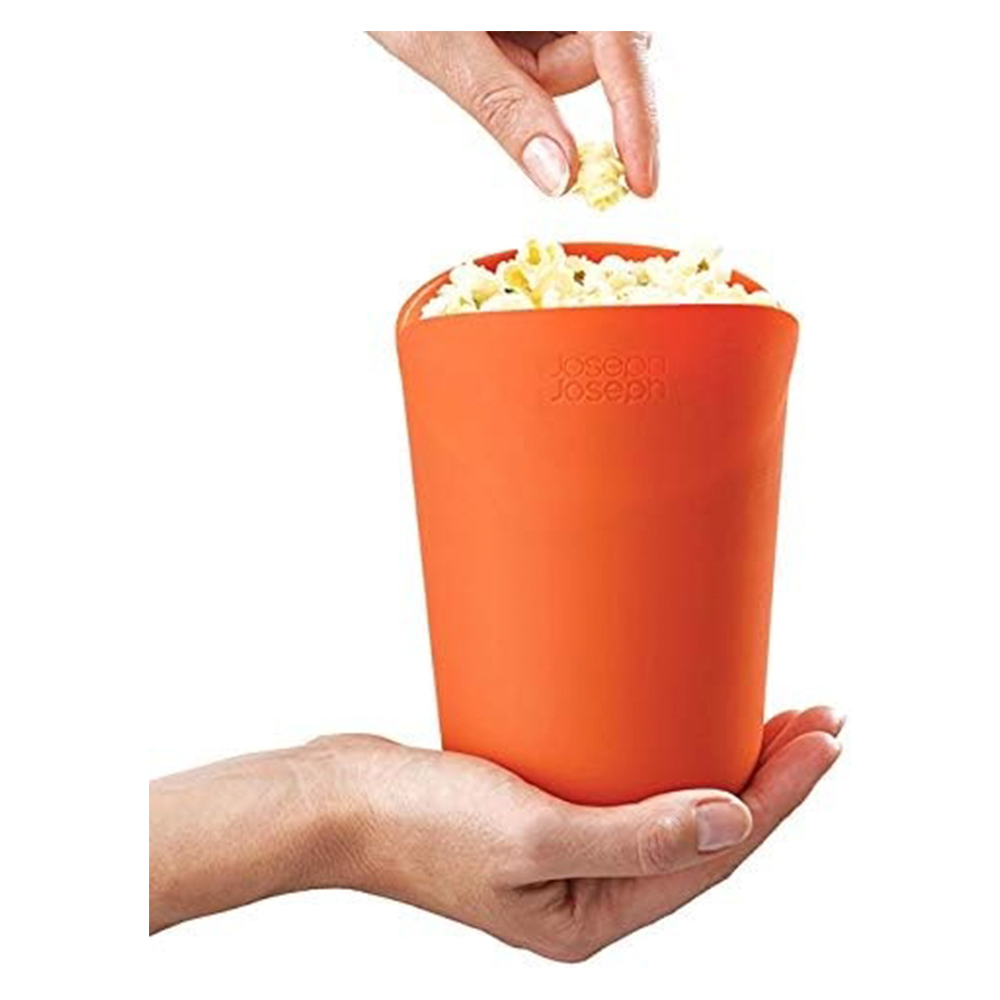 Mano sosteniendo una máquina de palomitas naranja llena de palomitas. Otra mano deja caer palomitas en el recipiente. Fondo blanco.