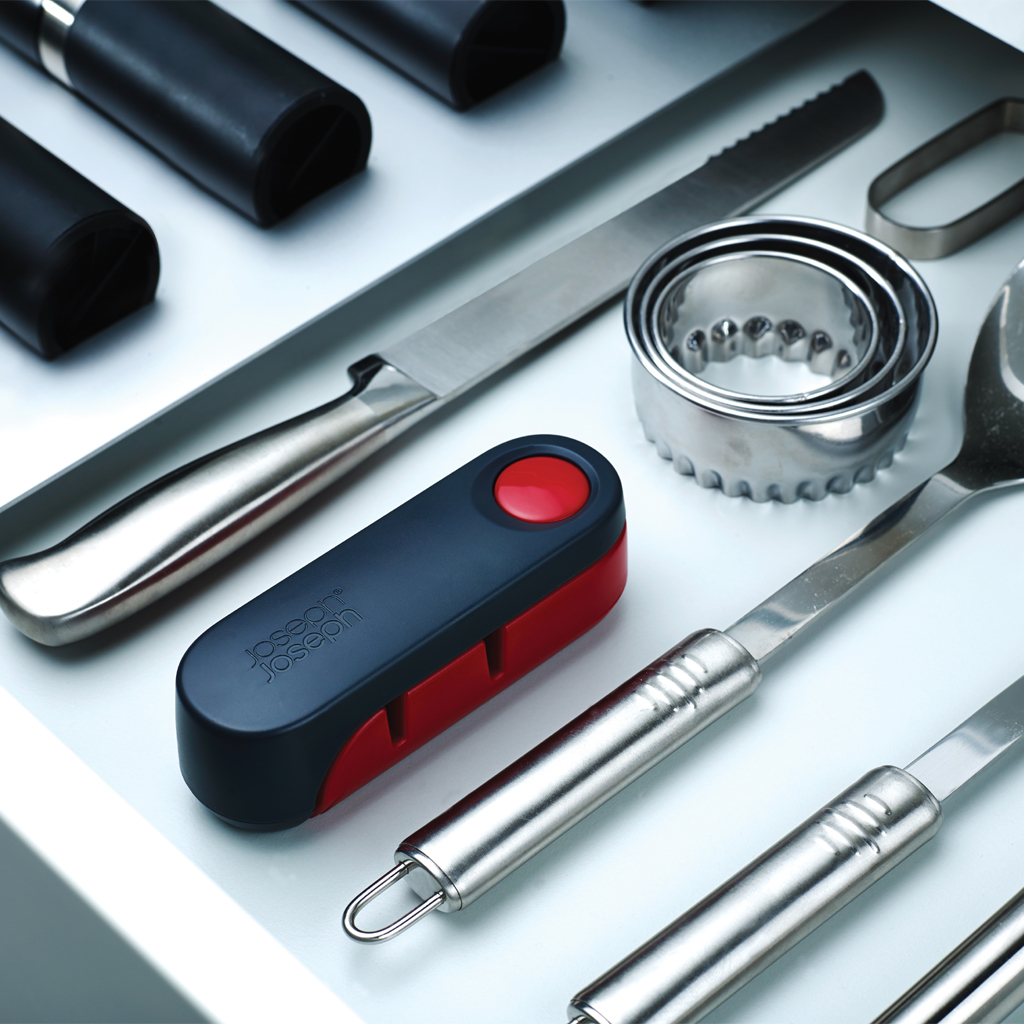 Utensilios de cocina sobre una superficie blanca: un cuchillo, un afilador, cortadores de galletas y cucharas. Colores: plata, negro y rojo.