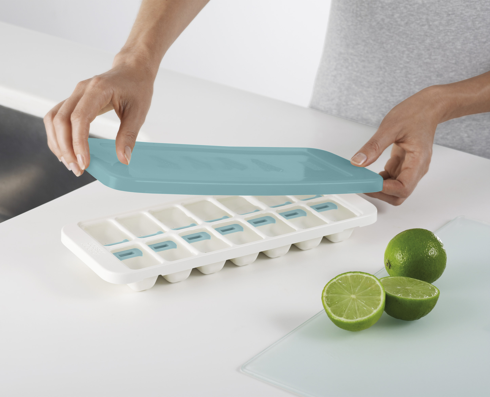 Manos sosteniendo una tapa azul claro sobre una cubitera blanca con detalles azules. Limas sobre una tabla de cortar.