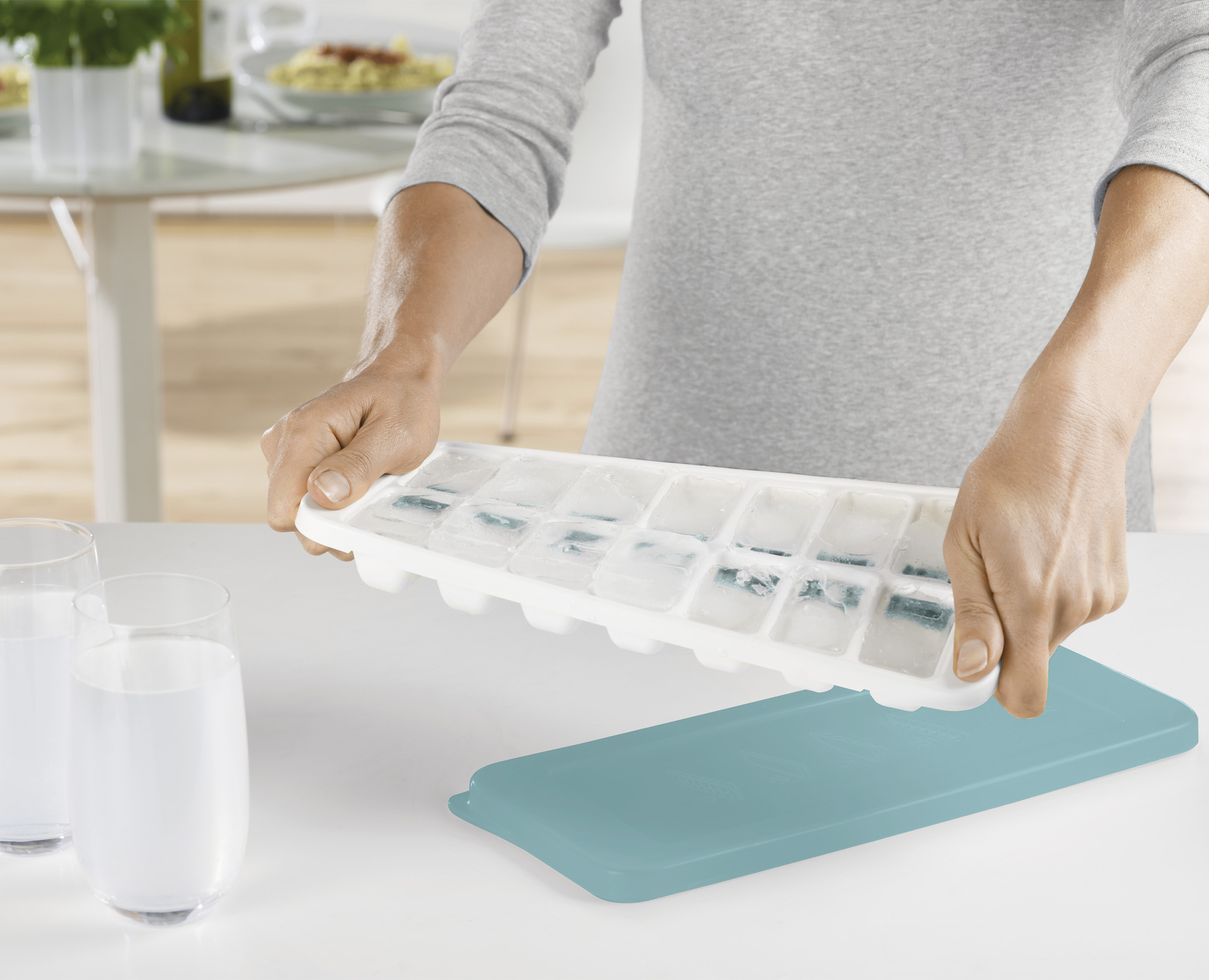Una persona sostiene una cubitera sobre una tapa verde azulado, con dos vasos de agua y una mesa con comida al fondo.