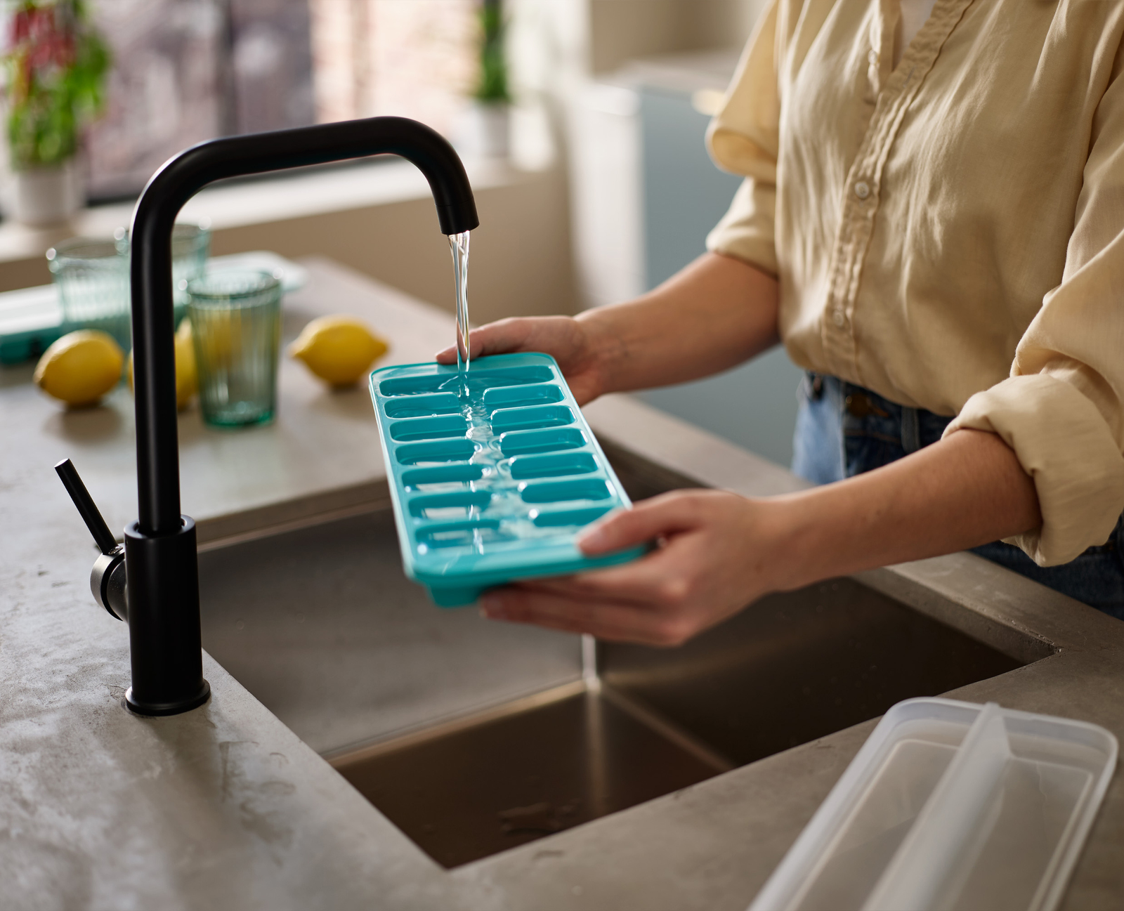 Una persona llena una cubitera azul con agua de un grifo negro. Hay limones y vasos en la encimera.