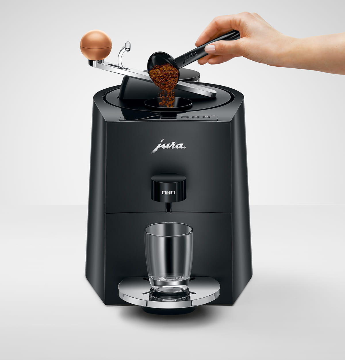 Eine Hand schüttet Kaffeepulver in eine schwarze Jura Kaffeemaschine. Ein Glas steht auf einem silbernen Tablett unter dem Auslauf.