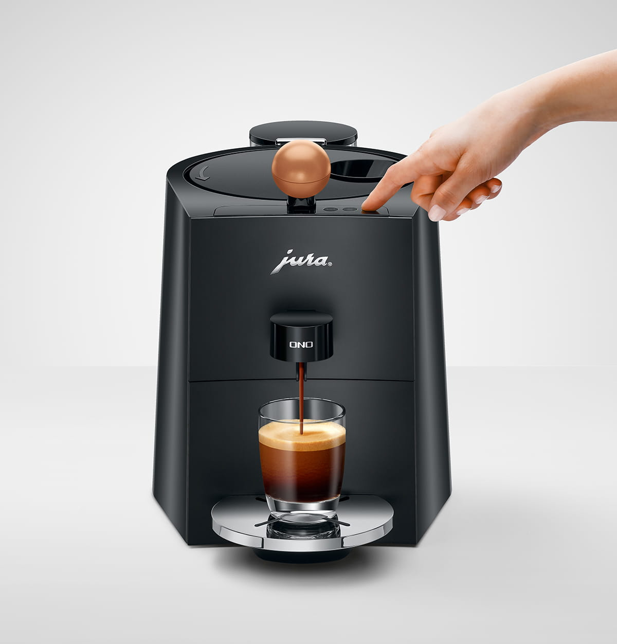 Een zwarte Jura ONO espressomachine schenkt koffie in een glas. Een hand drukt op een knop. Koperen bal bovenop.