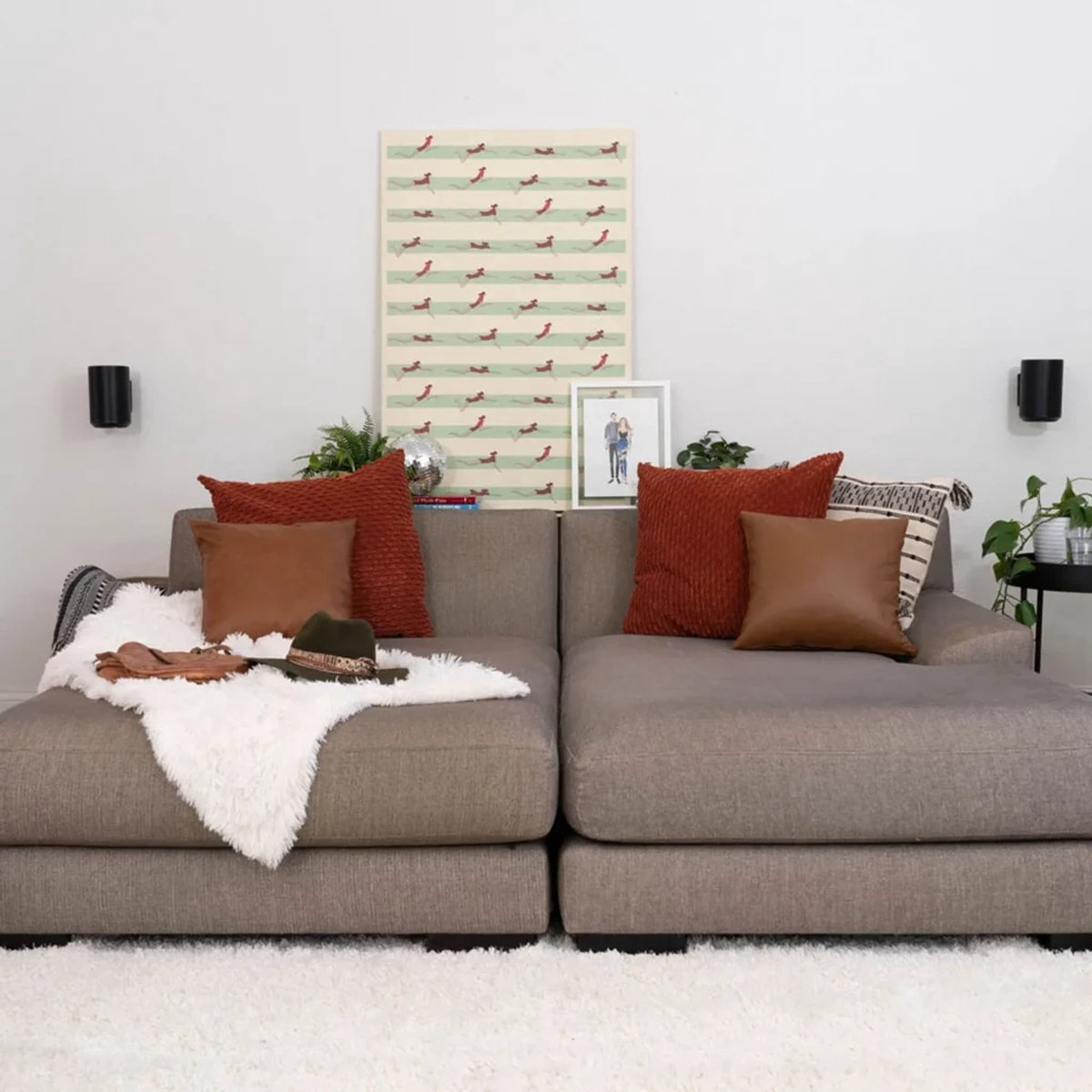A gray sectional sofa with pillows, a hat, and a painting of dogs. A white rug is on the floor, and a picture hangs.