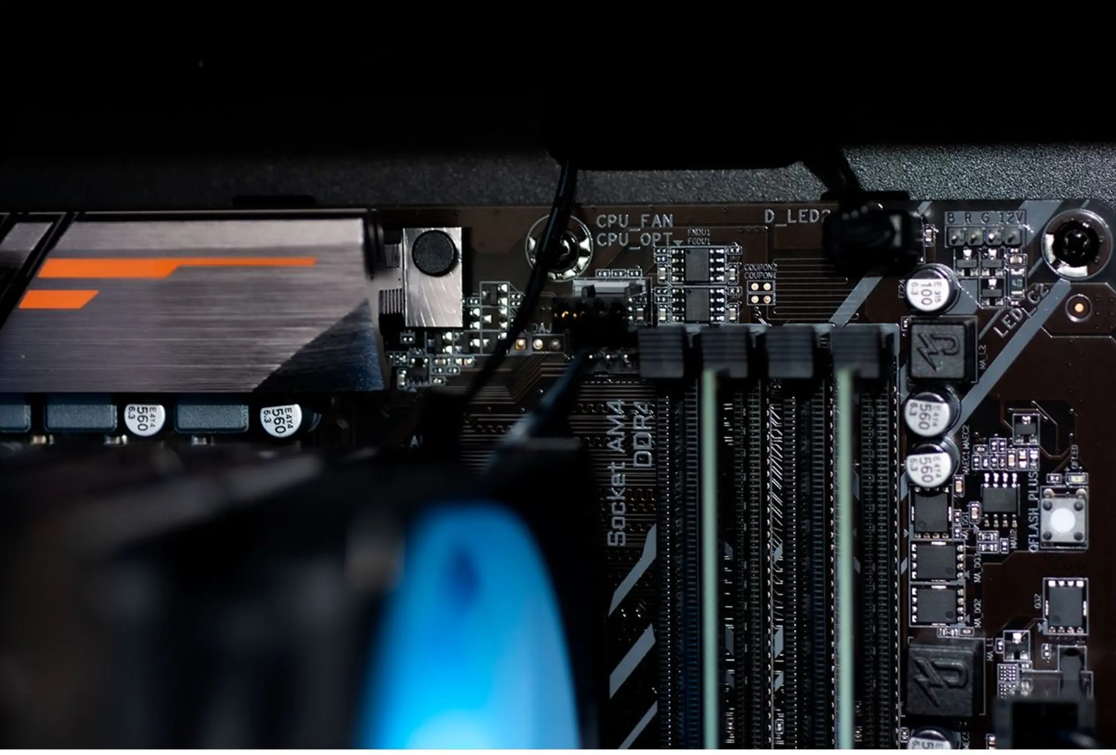 Close-up van een computer moederbord. Componenten omvatten RAM-sleuven, CPU-ventilator en diverse chips. Kleuren zijn zwart, zilver en oranje.