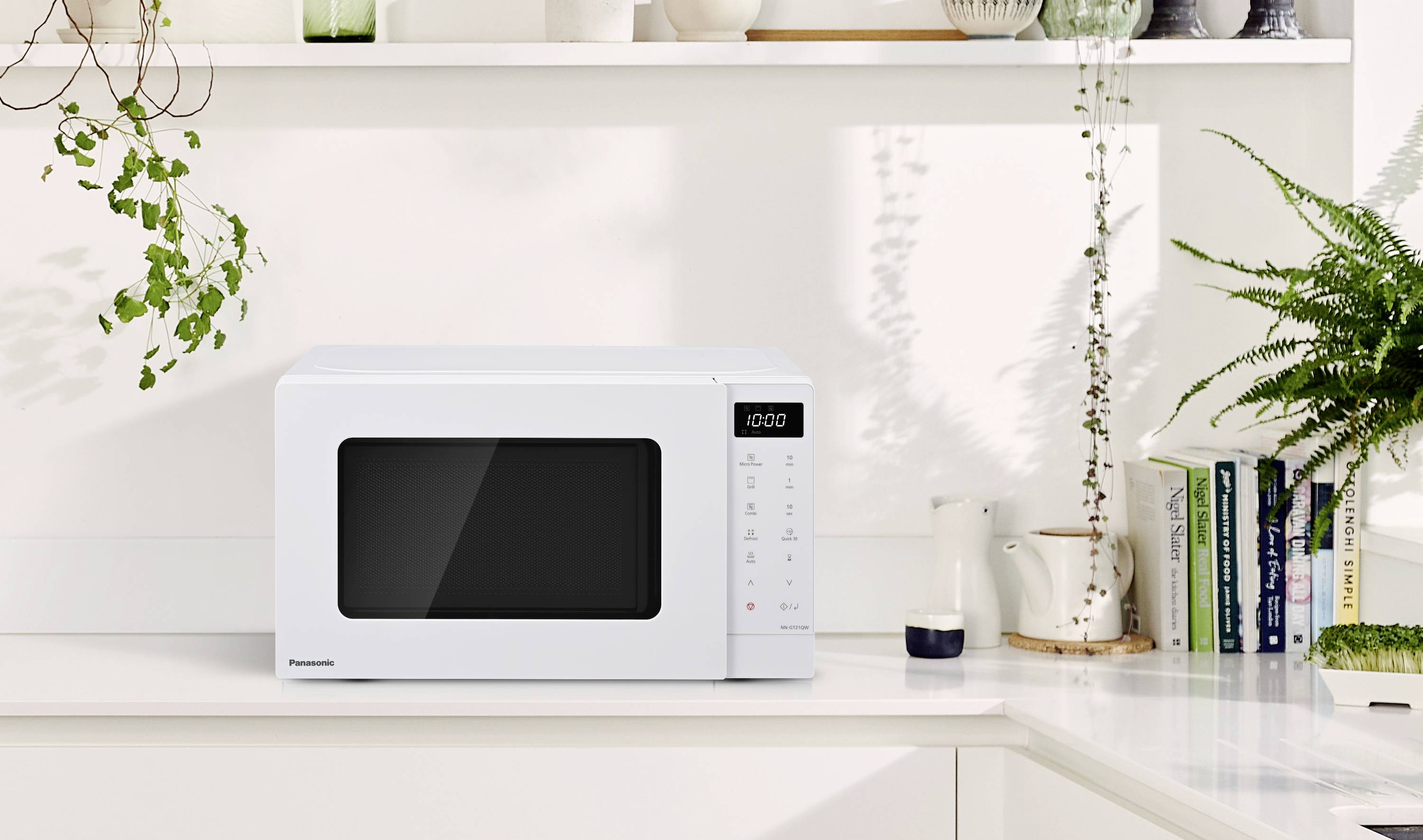 Un forno a microonde bianco con display digitale che mostra le 10:00, posto su un piano di lavoro bianco con piante e libri sullo sfondo.