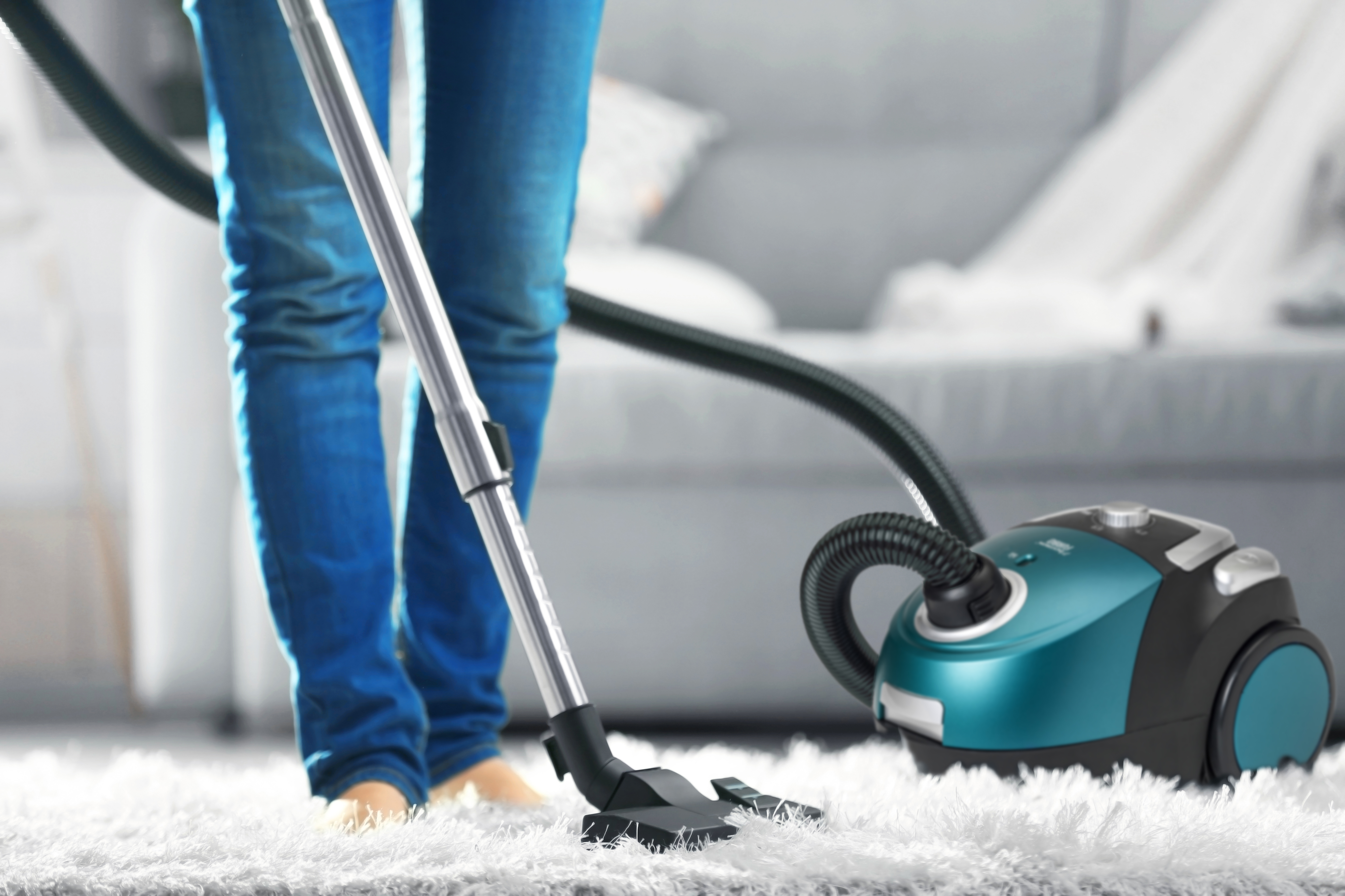Una persona aspira un tappeto bianco con un aspirapolvere turchese e nero. La persona indossa jeans.