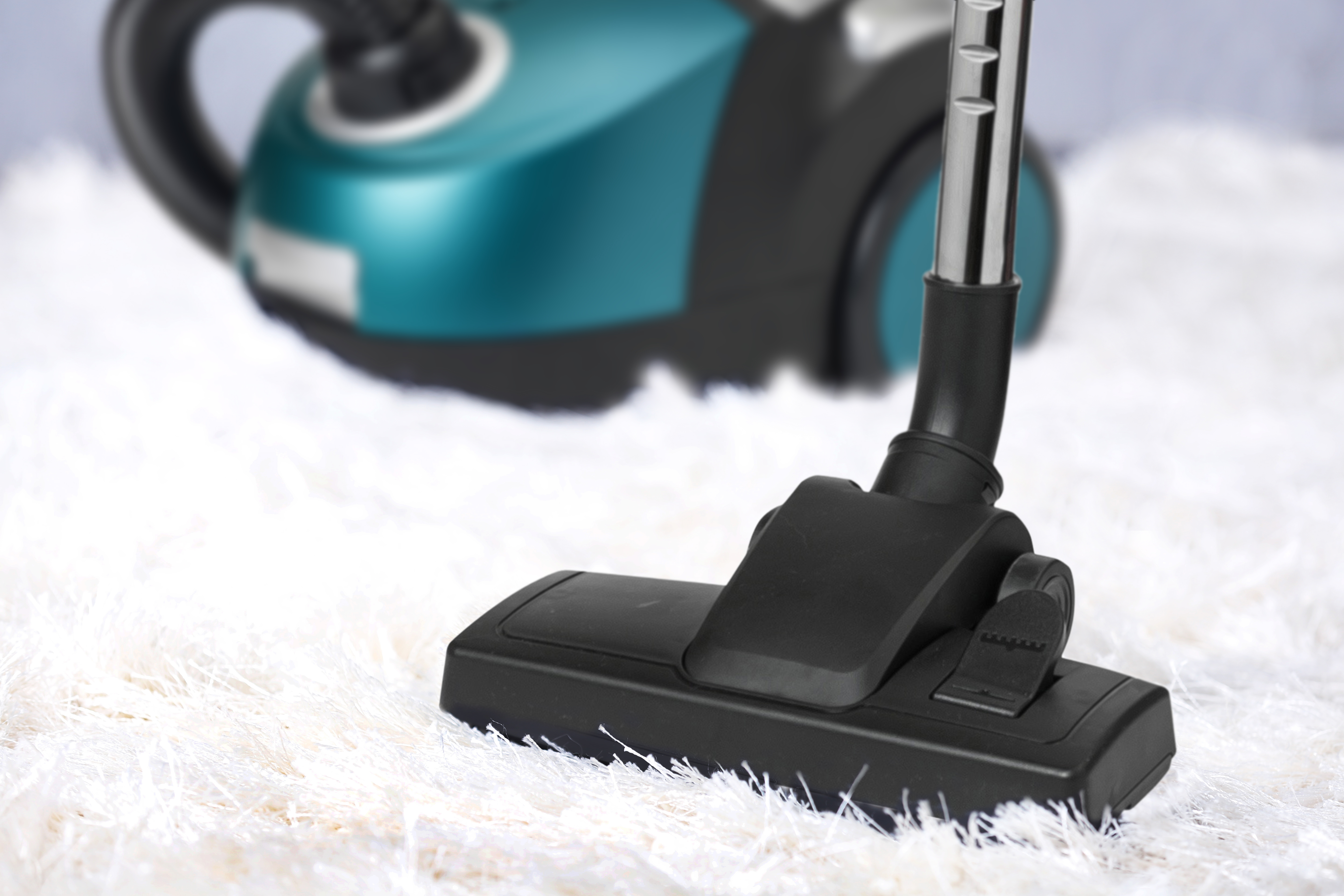 Una testa di aspirapolvere nera su un tappeto bianco e soffice. Sullo sfondo, è visibile un'aspirapolvere verde acqua e nera.