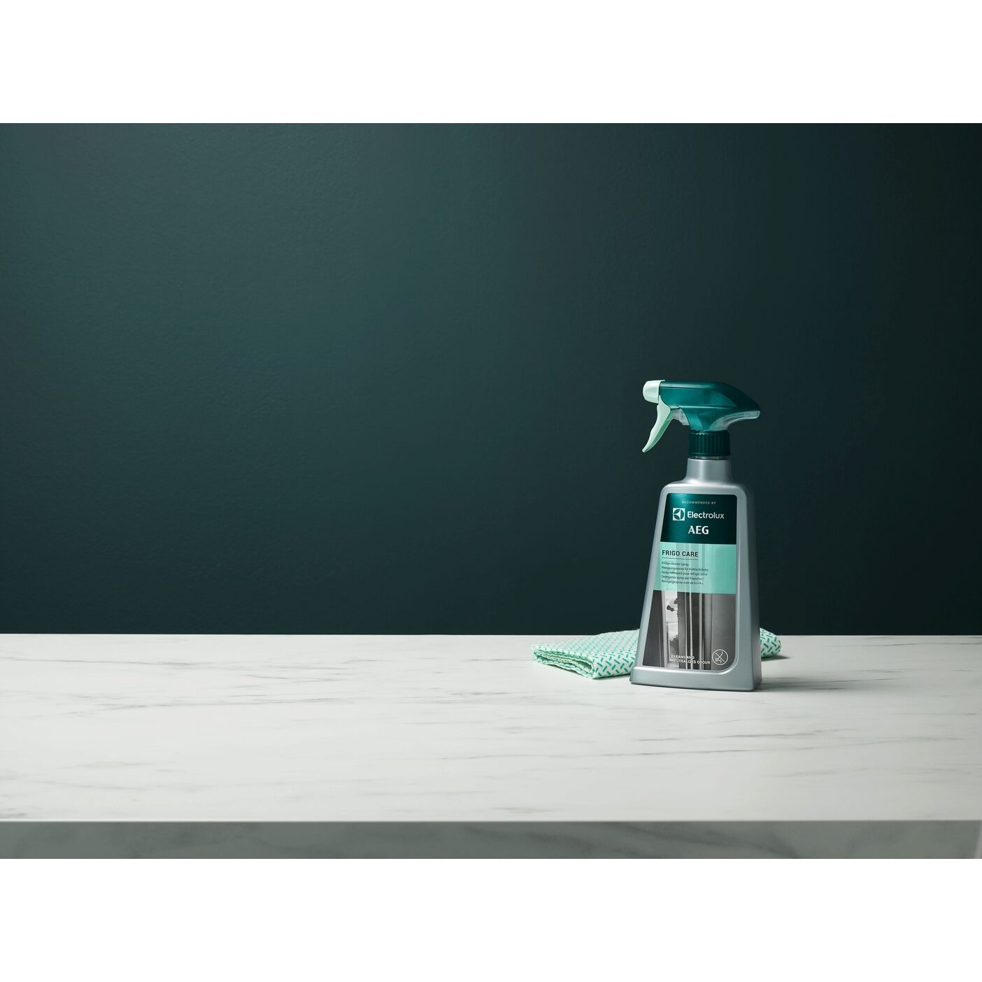 Una bottiglia di spray AEG Frigo Care su una superficie di marmo, con un panno verde. Parete verde scuro sullo sfondo.