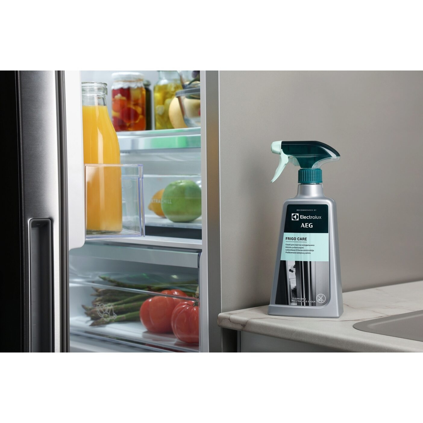 Una porta del frigorifero è aperta. All'interno ci sono verdure e succo. Accanto c'è una bottiglia di spray detergente AEG Frigo Care.