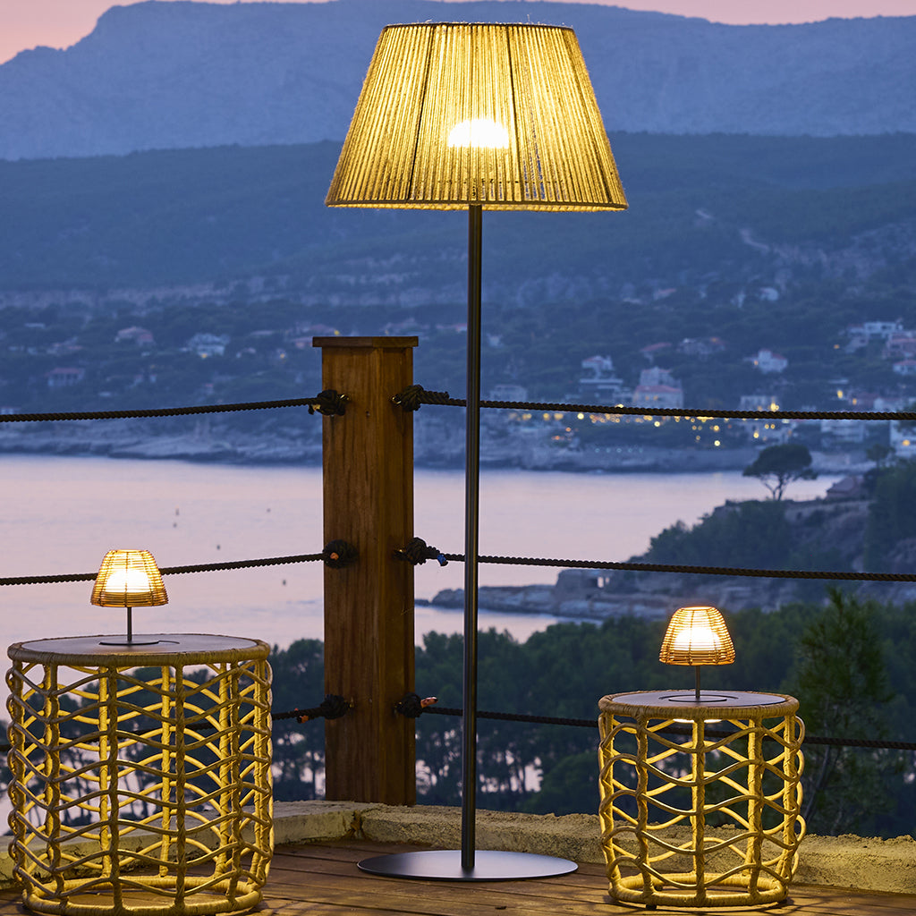 Außenszene mit Lampen auf Korbtischen, einer hohen Lampe und Meerblick im Hintergrund bei Dämmerung.