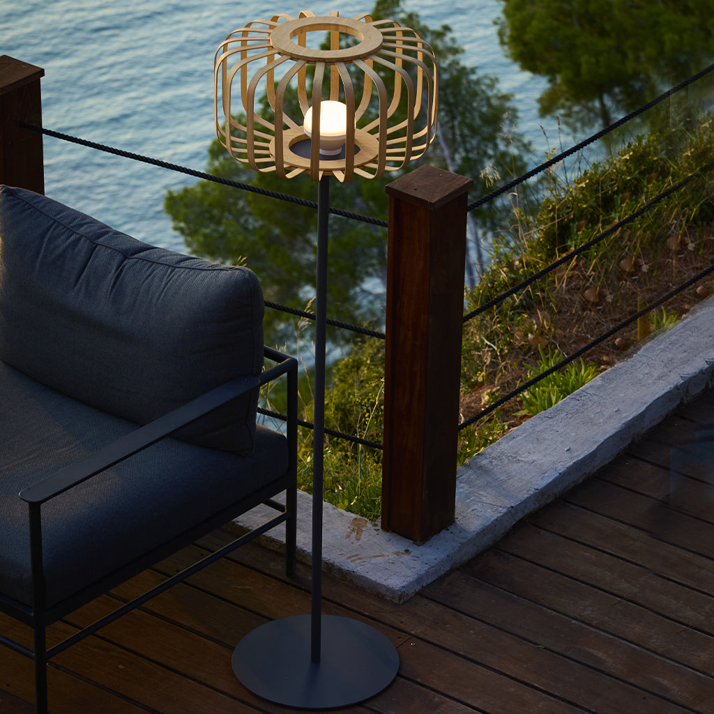 Eine dekorative Lampe mit Holzschatten steht auf einem Deck. Ein dunkles Sofa steht daneben, mit Blick auf das Meer im Hintergrund.