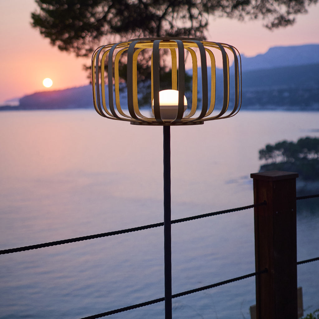 Eine beleuchtete Lampe mit käfigartigem Lampenschirm steht auf einer Stange. Sonnenuntergang über dem Meer im Hintergrund. Ein Zaun ist im Vordergrund.