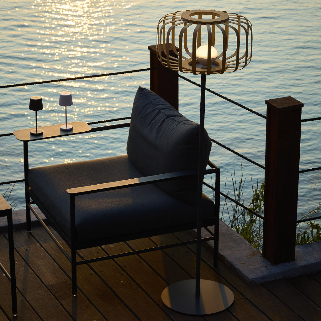 Eine moderne Szene im Freien mit einem Stuhl, Lampen und Meer im Hintergrund. Die Beleuchtung ist warm. Ein Holzdeck und ein Geländer sind sichtbar.