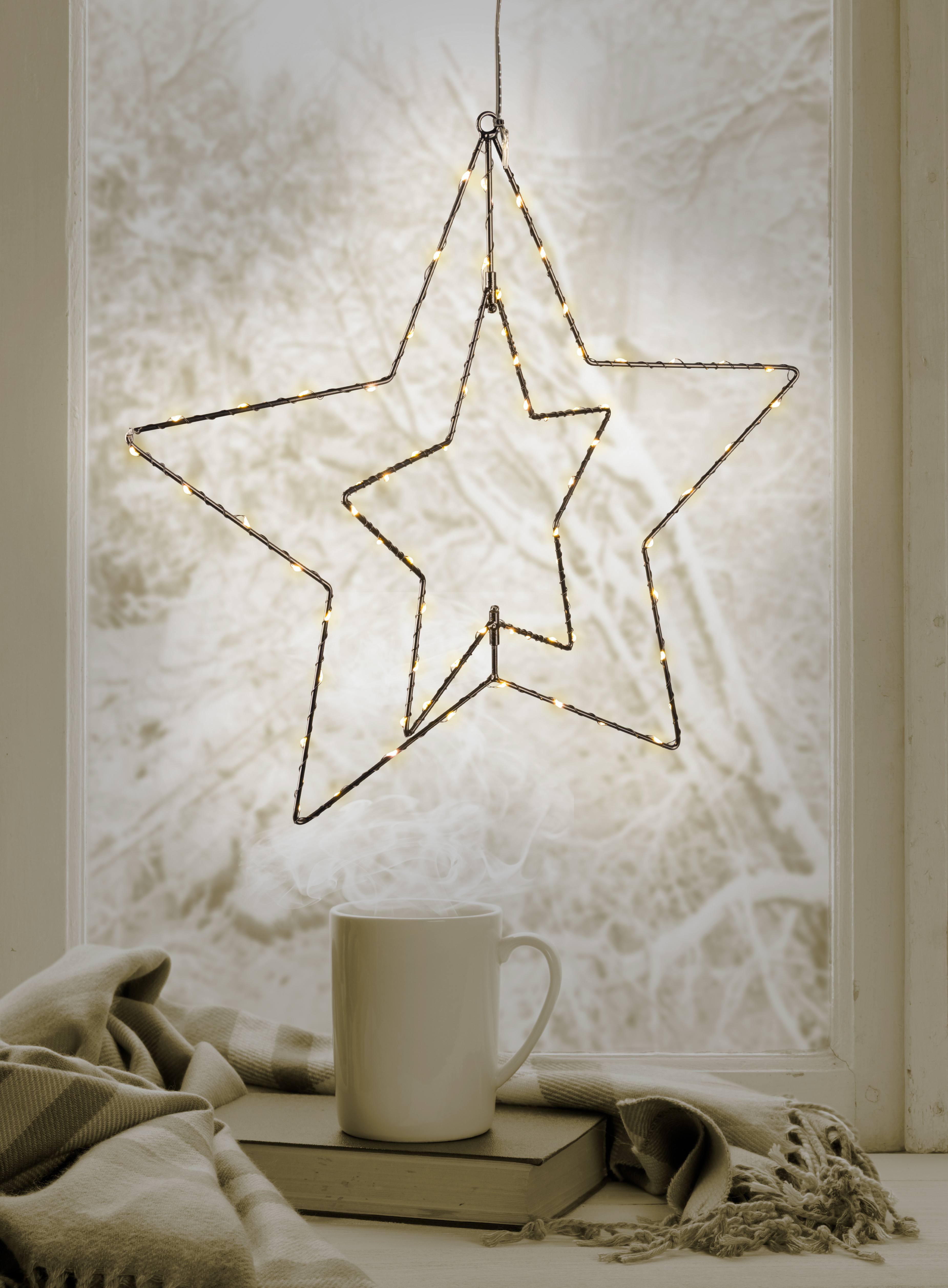 Ein sternförmiges Licht hängt vor einem Fenster. Ein Kaffeebecher steht auf einem Buch in der Nähe einer Decke.