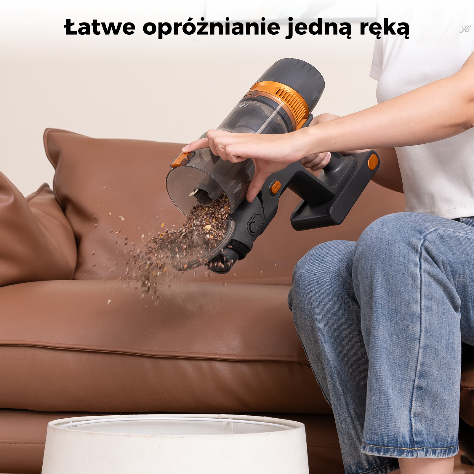 Osoba opróżnia odkurzacz z brązową kanapą w tle. Odkurzacz jest czarny i pomarańczowy.