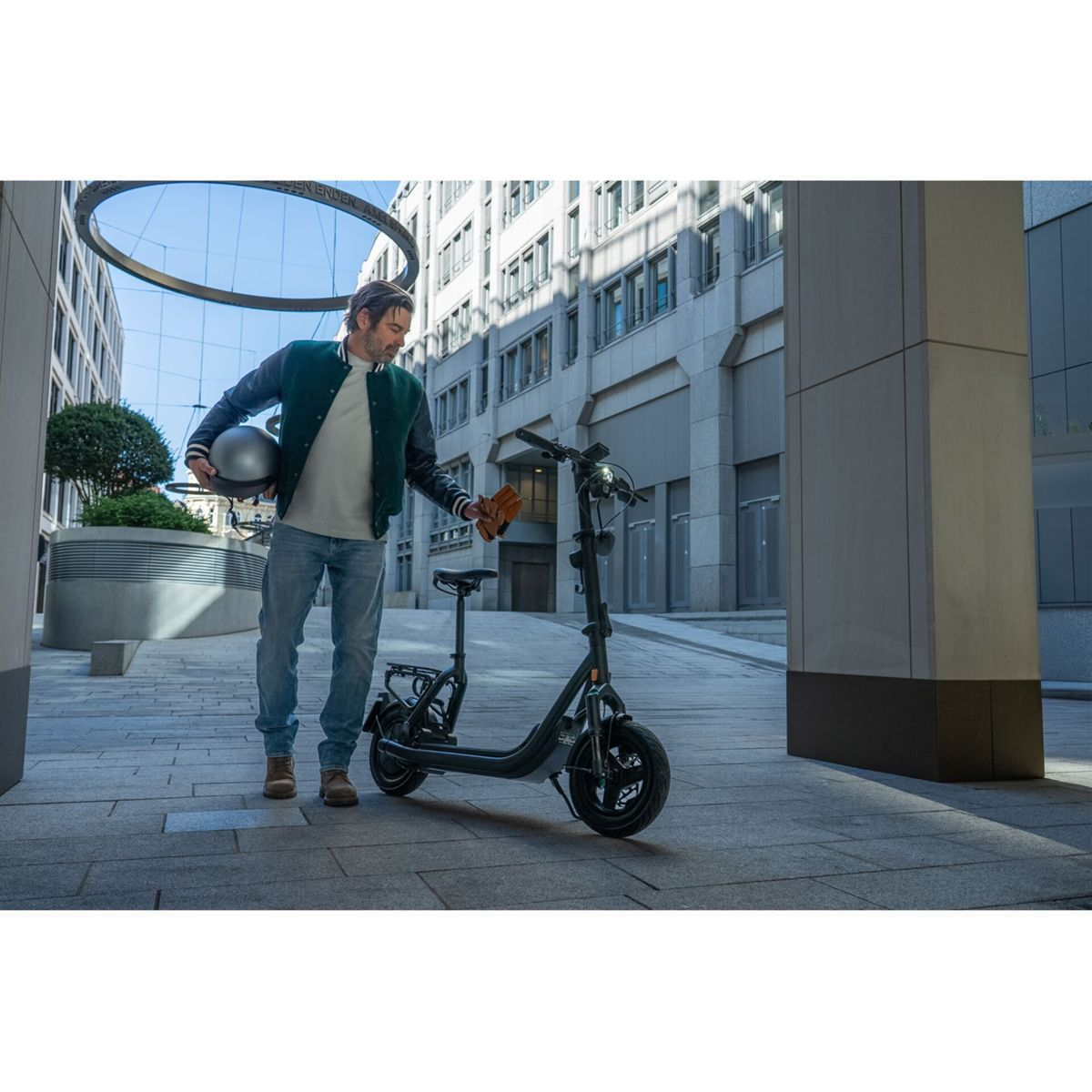 Ein Mann in einer grünen Jacke hält einen Helm und einen Handschuh und steht in einer Stadt neben einem schwarzen Elektroroller.