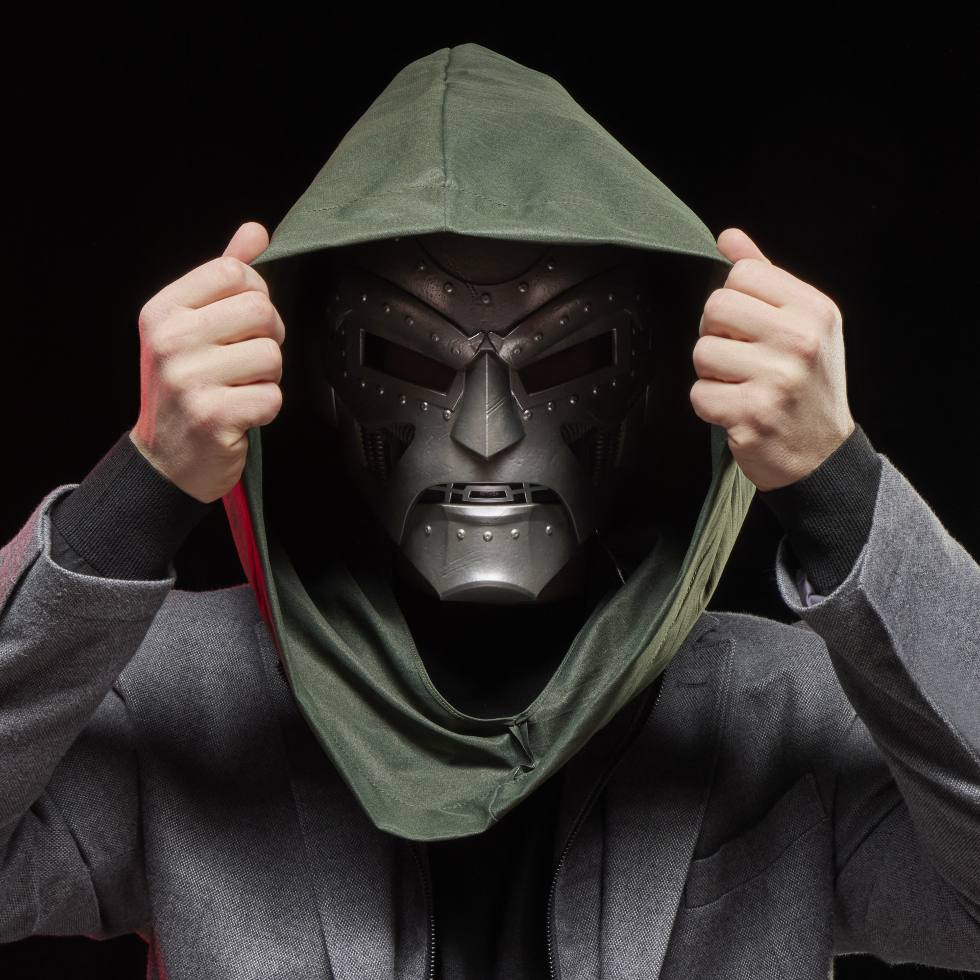Doktor Doom maskesi ve yeşil kapüşonlu kişi, eller kapüşonu çekiyor. Karanlık arka plan.