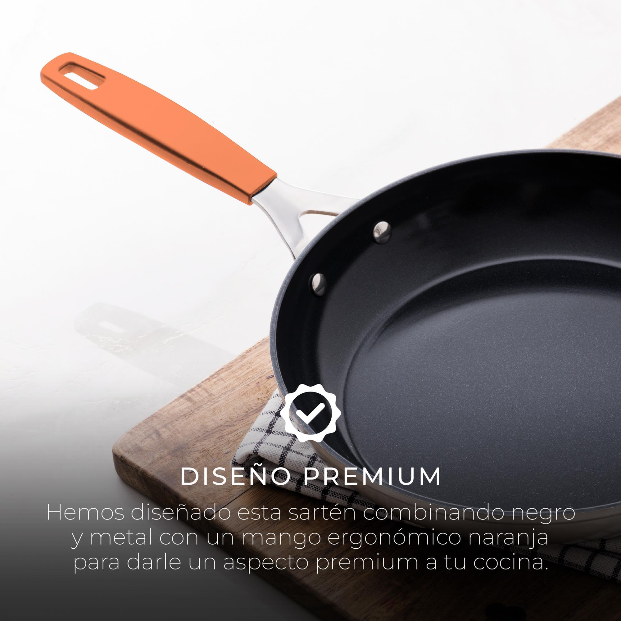 Una padella nera con manico arancione poggia su un tagliere di legno con un panno a scacchi. Sfondo bianco.