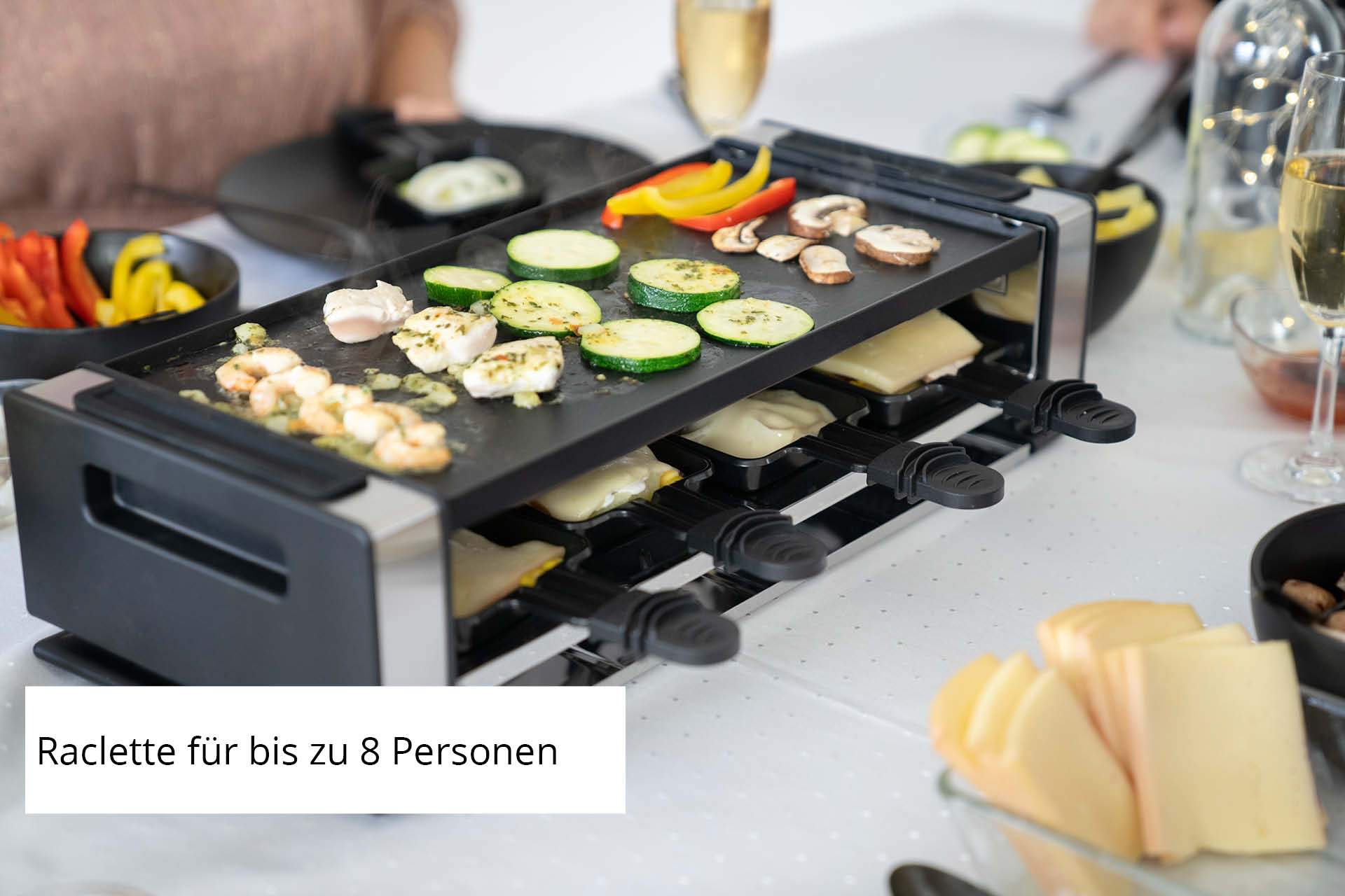 Ein Raclette-Grill mit Gemüse, Pilzen und Garnelen. Käse schmilzt darunter. Weingläser und Schalen sind vorhanden.