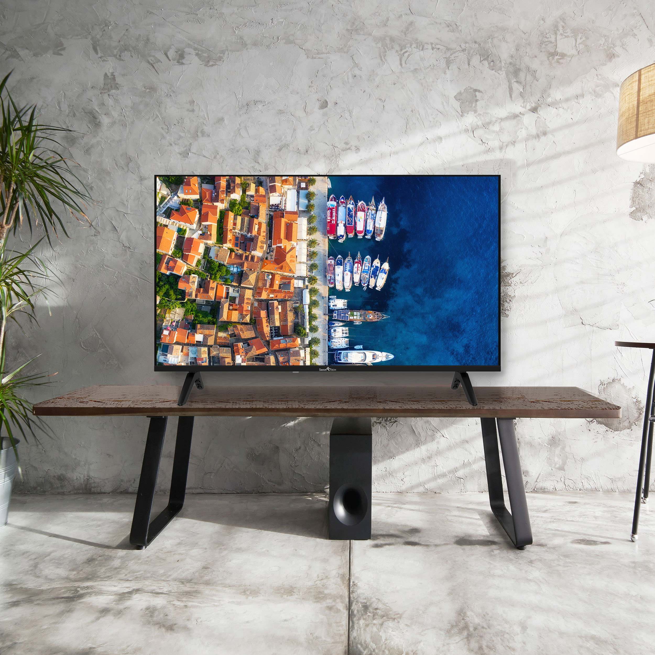 El televisor muestra un pueblo costero con barcos. Está sobre una mesa de madera con patas negras y un subwoofer debajo.