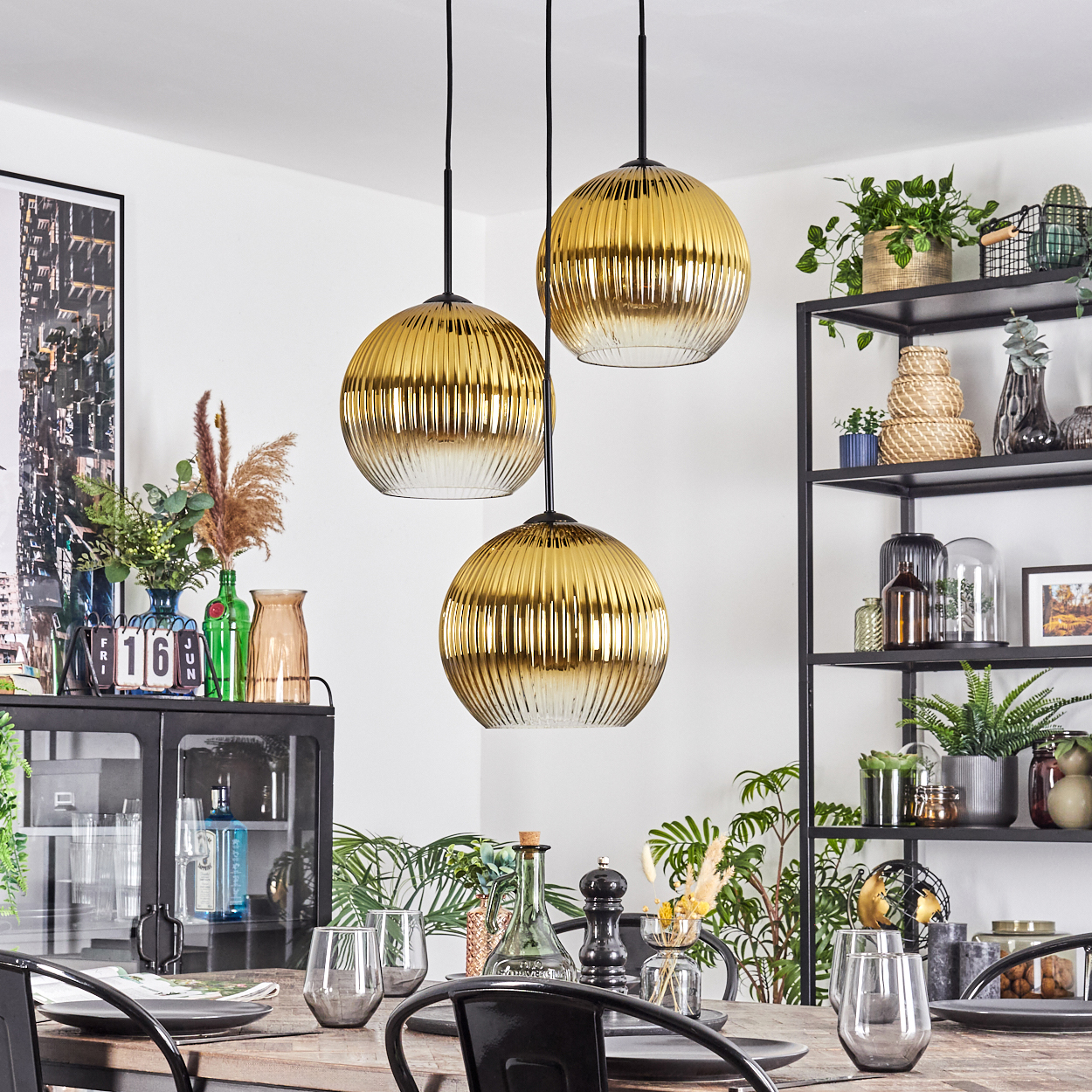 Esszimmer mit drei goldenen Pendelleuchten, einem gedeckten Tisch und einem schwarzen Regal mit Pflanzen und Dekoration.