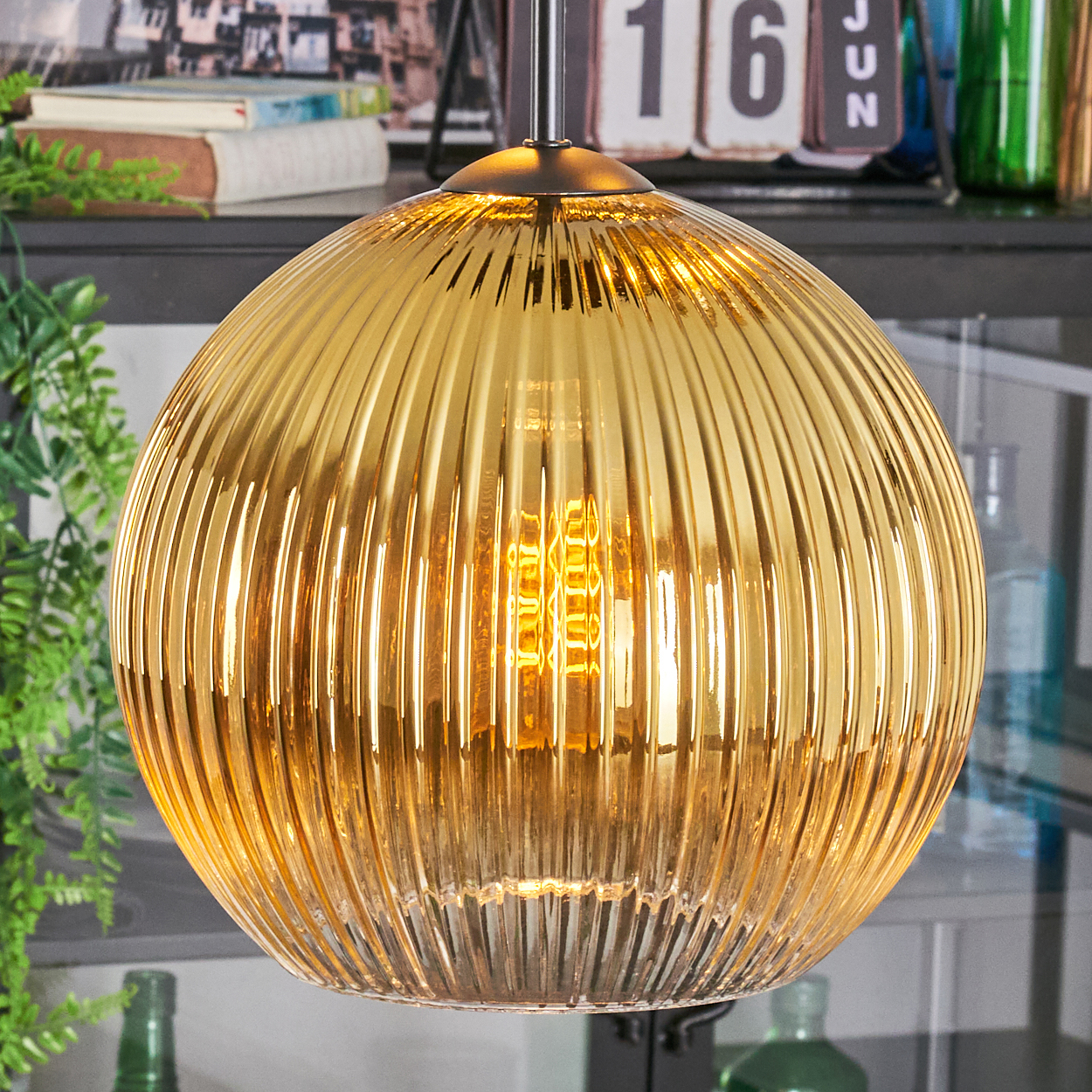 Eine goldene, gerippte Glas-Pendelleuchte mit leuchtender Glühbirne hängt. Der Hintergrund umfasst Bücher, Kalender und grüne Pflanze.