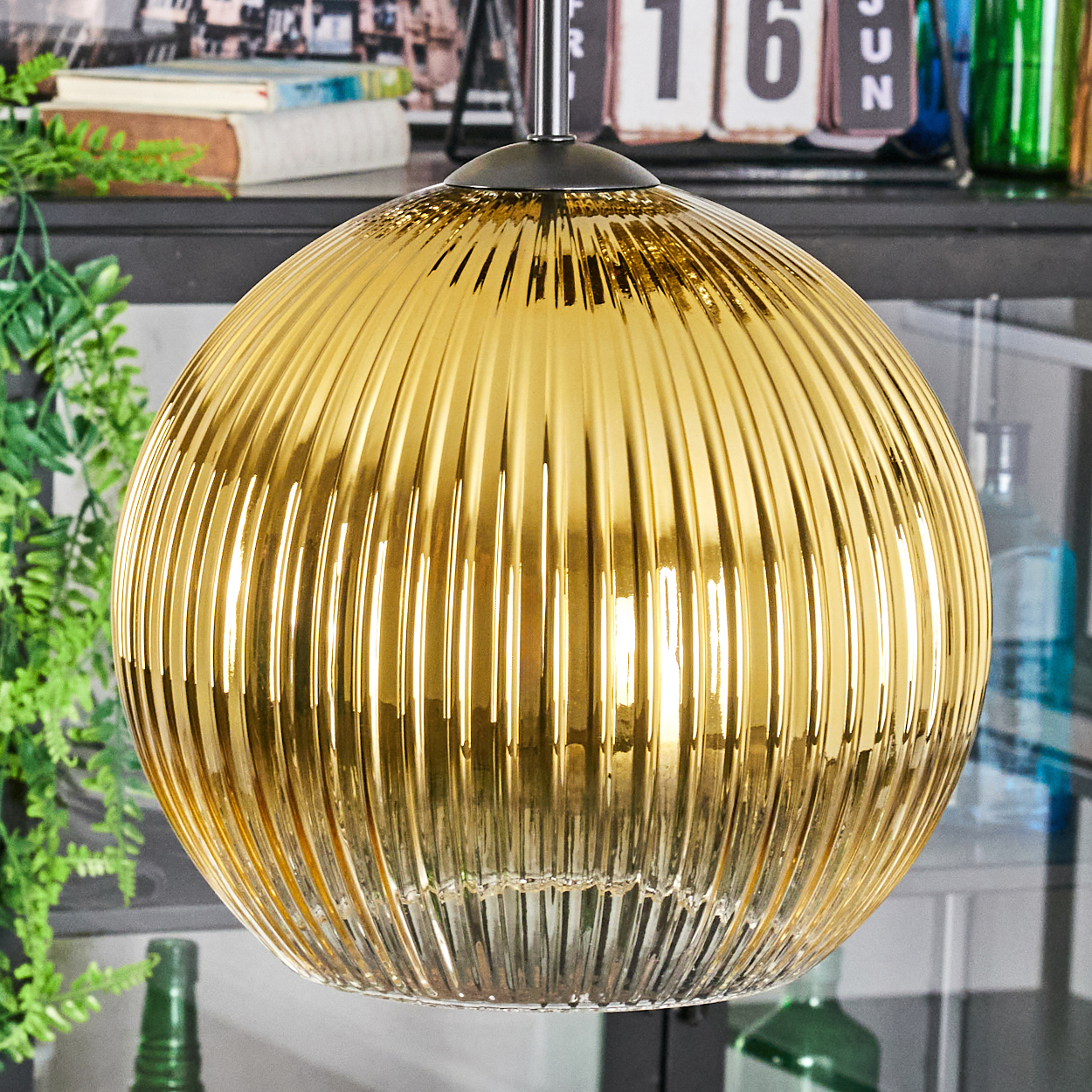Eine goldene, gerippte Glas-Pendelleuchte hängt. Der Hintergrund umfasst Grün, Bücher und einen Kalender. Unten ist eine grüne Flasche zu sehen.