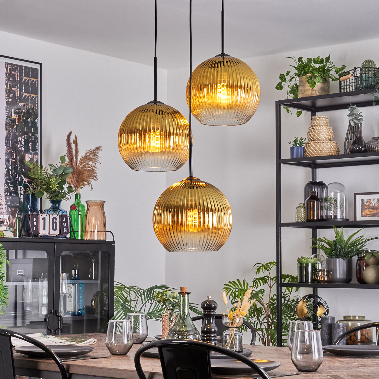 Esszimmer mit drei goldenen Pendelleuchten. Tisch mit Gläsern und Tellern. Regale mit Pflanzen und Dekor.