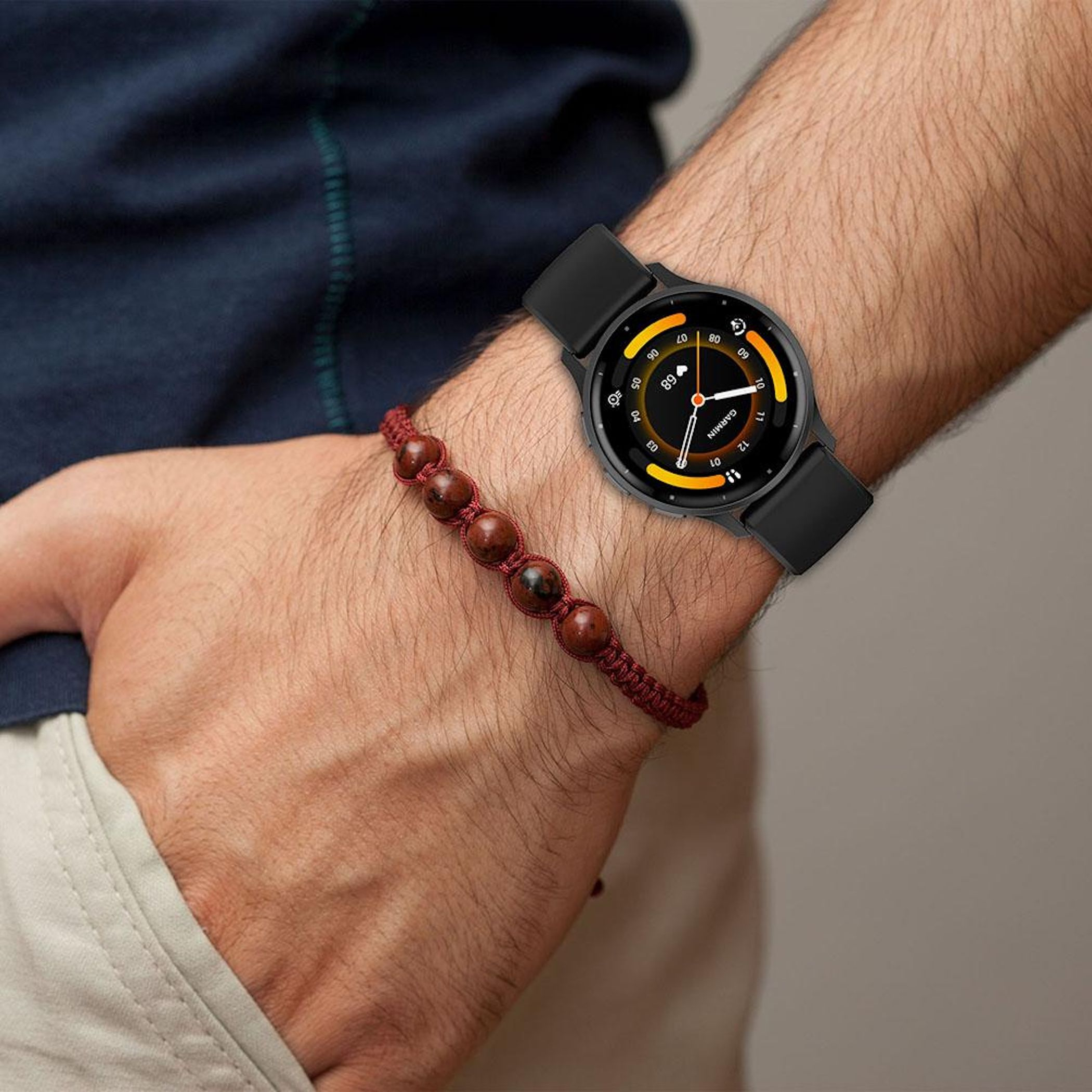 Ein Handgelenk mit einer schwarzen Smartwatch und einem roten Perlenarmband. Im Hintergrund ist ein dunkelblaues Hemd zu sehen.
