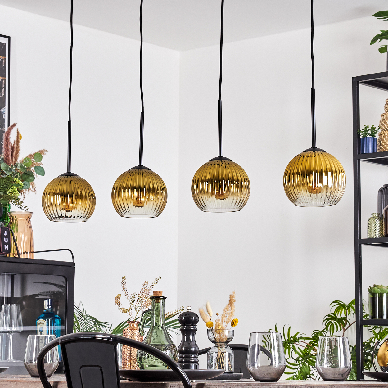 Vier goldene Pendelleuchten hängen über einem Esstisch, der mit Gläsern und Dekor gedeckt ist. Pflanzen und Regale sind im Hintergrund zu sehen.